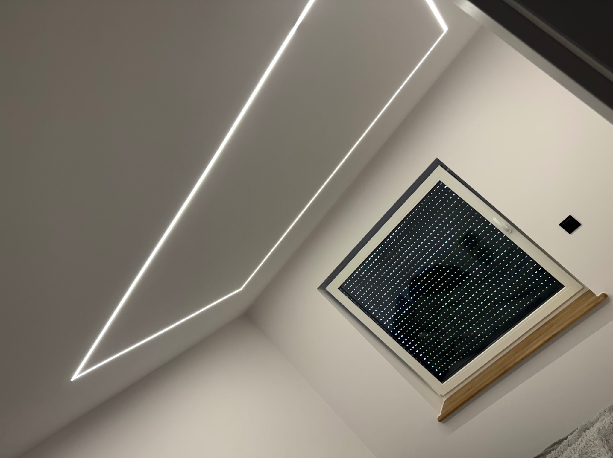 Nowoczesne oświetlenie LED w kształcie geometrycznym na suficie poddasza, obok okna dachowego z roletami. Minimalistyczny design wnętrza.