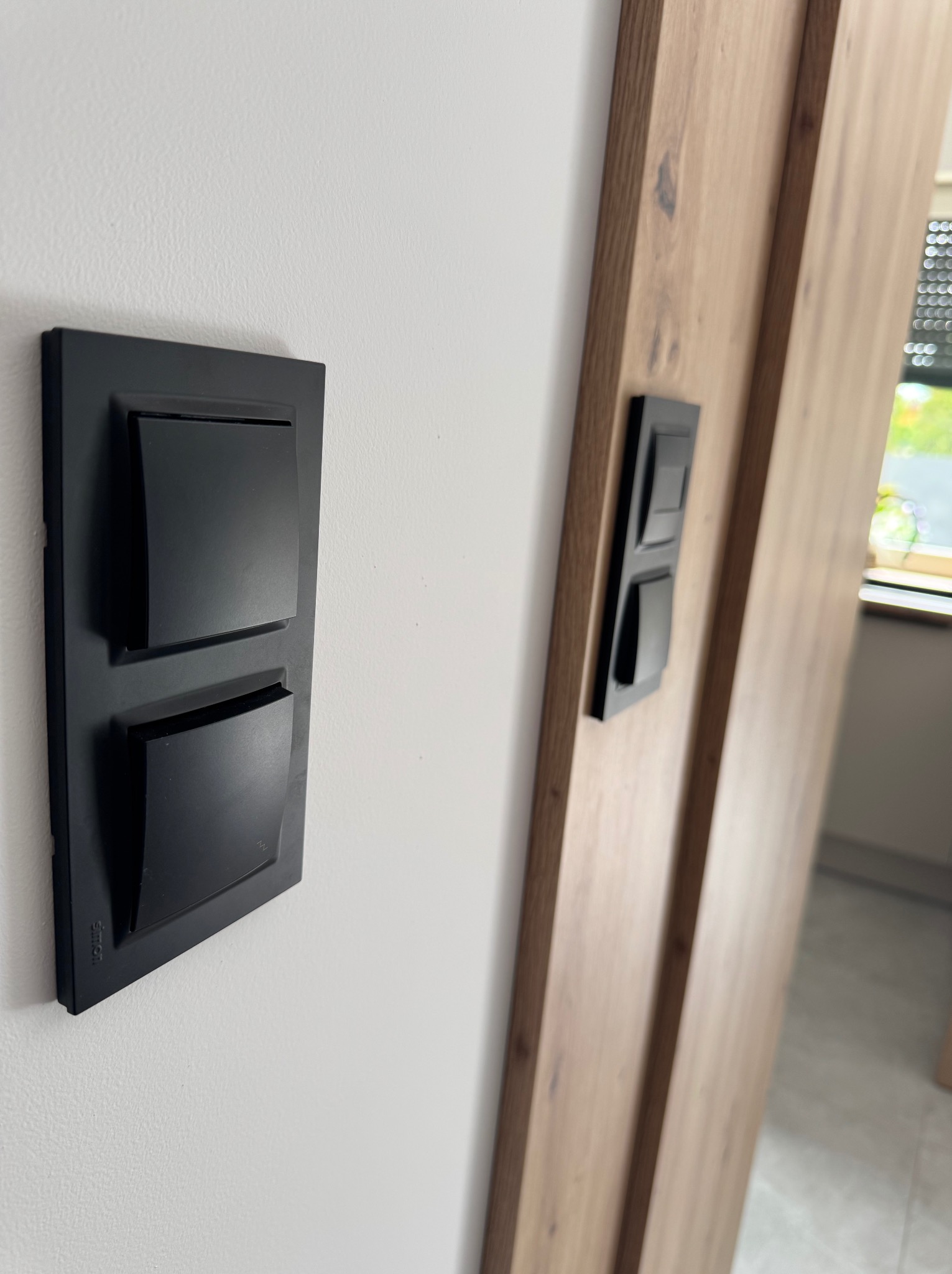 Nowoczesny, czarny podwójny włącznik światła Simon na białej ścianie, obok drewnianej ościeżnicy z drugim włącznikiem. Minimalistyczny design, wnętrze domu.