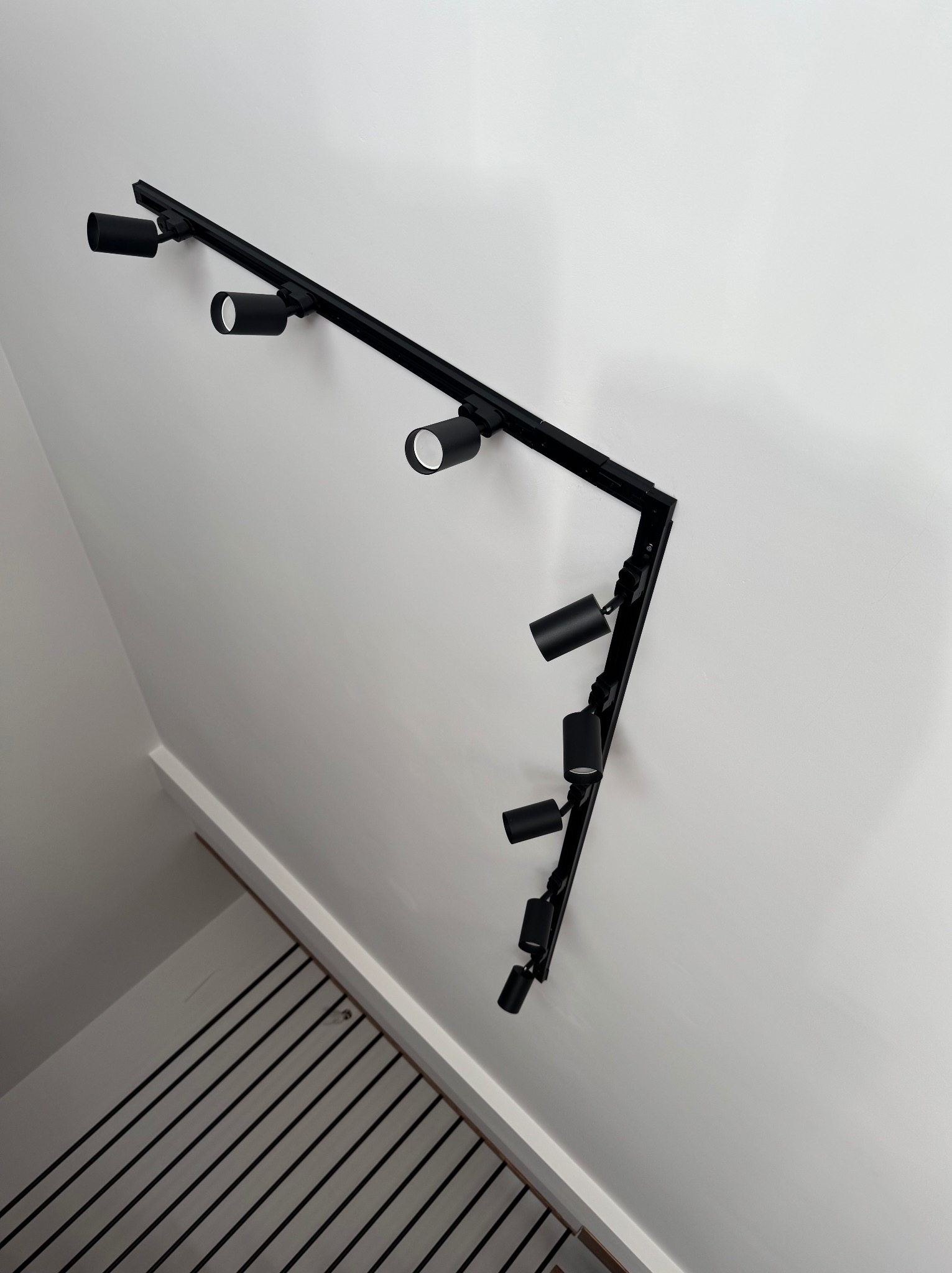 Czarna, szynowa instalacja oświetleniowa z reflektorami, zamontowana na białej ścianie w rogu pomieszczenia. Widok z dołu, fragment schodów w tle.