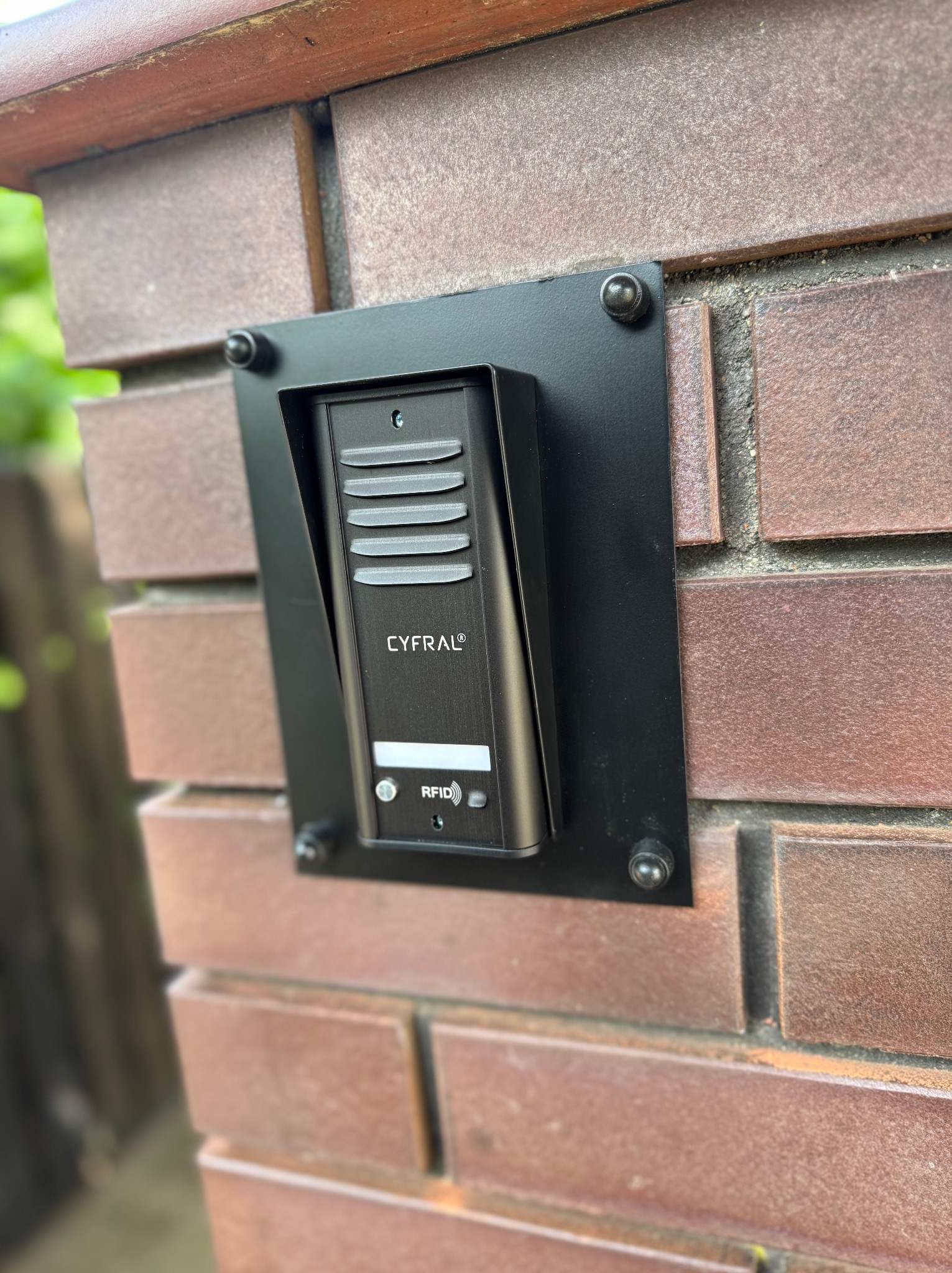 Czarny wideodomofon Cyfral z czytnikiem RFID zamontowany na ceglanym słupku ogrodzenia, widoczne śruby mocujące i fragment zieleni w tle.