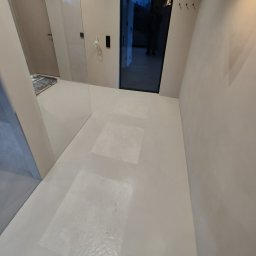 PROFITECH - Borys Tustanovskyi - Jasne, minimalistyczne wnętrze z gładką, białą posadzką, na której widoczne są kwadratowe plamy po zabezpieczeniu podłoża folią malarską, drzwi wejściowe z czarną ramą i widocznym odbiciem sylwetki...