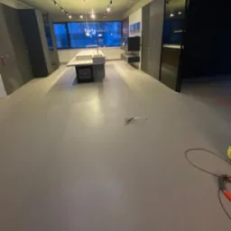 Wnętrze nowocześnie wykończonej kuchni z szarą, gładką podłogą, widok na okno i minimalistyczną zabudowę kuchenną, w tle widoczne narzędzia i kable.