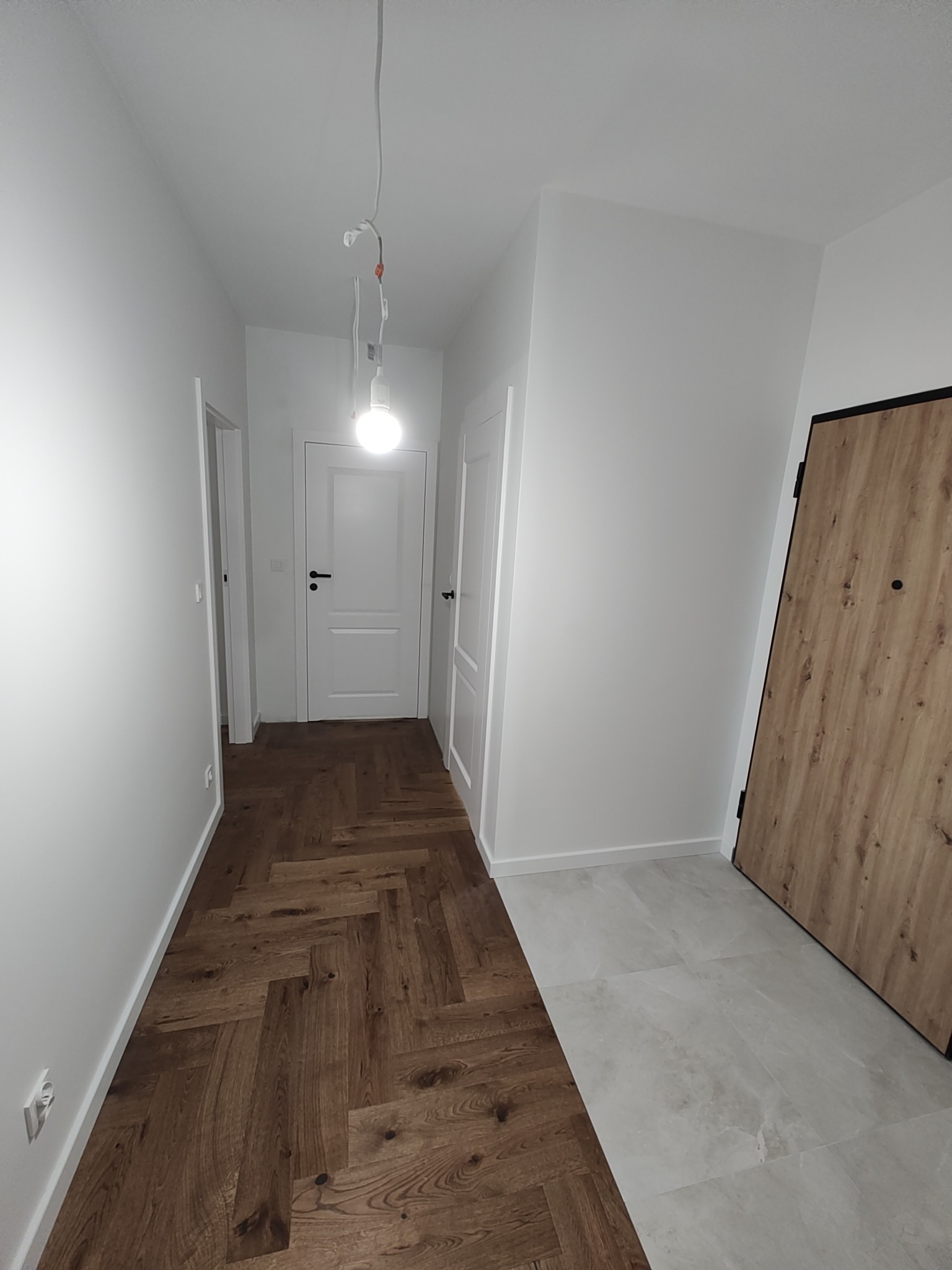 Przedpokój po remoncie z dwiema różnymi podłogami: parkiet w jodełkę i jasne płytki, białe ściany i drzwi wewnętrzne, oraz drewnopodobne drzwi wejściowe z czarną ramą.