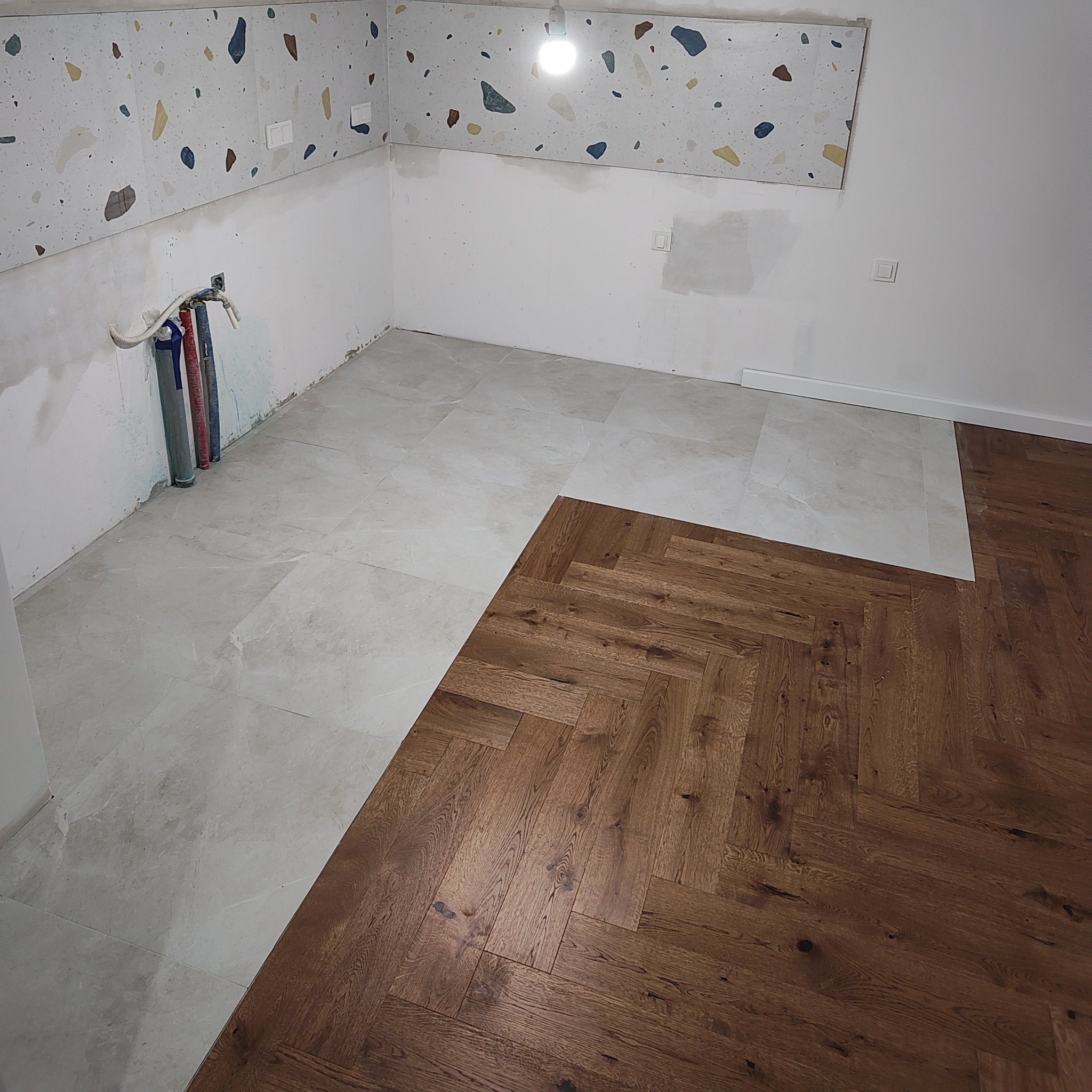 Podłoga w trakcie układania: parkiet w jodełkę łączy się z szarymi płytkami podłogowymi, ściana z kolorowym wzorem lastryko w tle, widoczne rury instalacyjne.