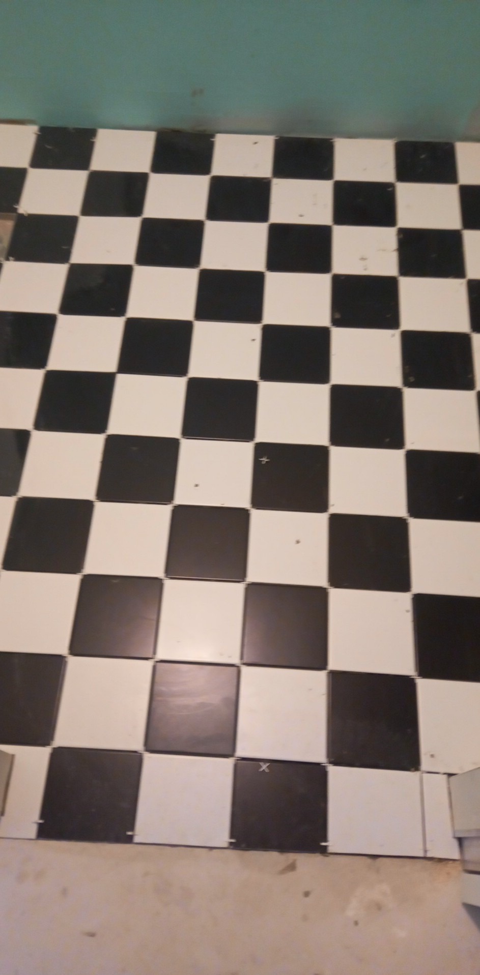 Ułożona podłoga w czarno-białą szachownicę z płytek ceramicznych, widoczne niedociągnięcia przy ścianie i resztki kleju.
