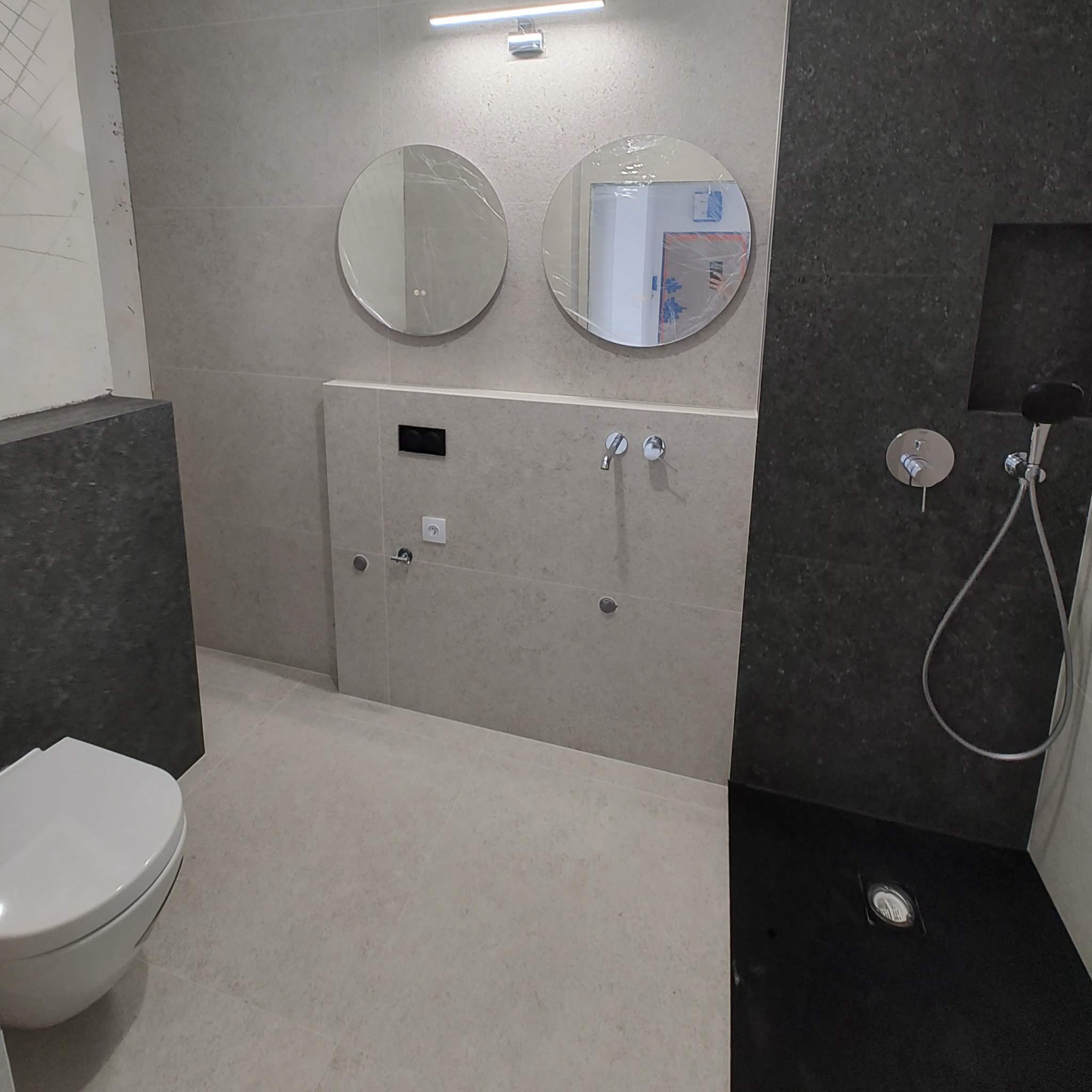Nowoczesna łazienka z szarymi płytkami, okrągłymi lustrami z folią ochronną, czarną kabiną prysznicową typu walk-in i podwieszaną toaletą, oświetlona górnym światłem.