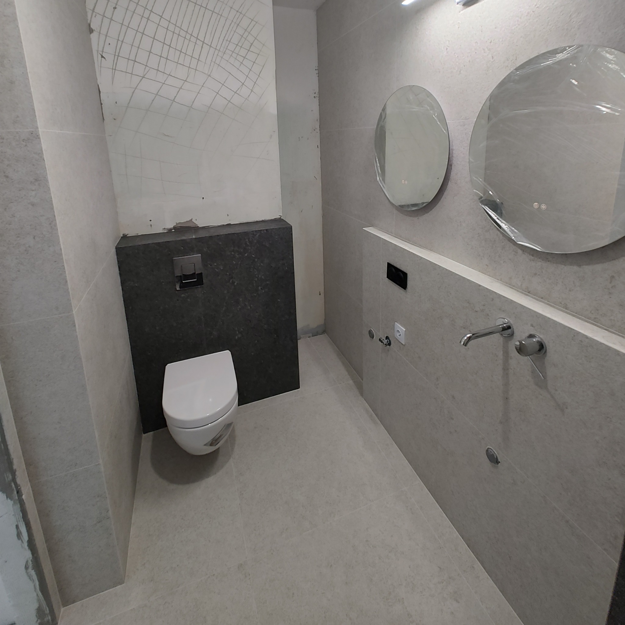 Nowoczesna łazienka w trakcie wykańczania, z podtynkową spłuczką WC, umywalką z baterią i okrągłymi lustrami z podświetleniem LED, zabezpieczonymi folią ochronną, widoczne ślady po szpachlowaniu...