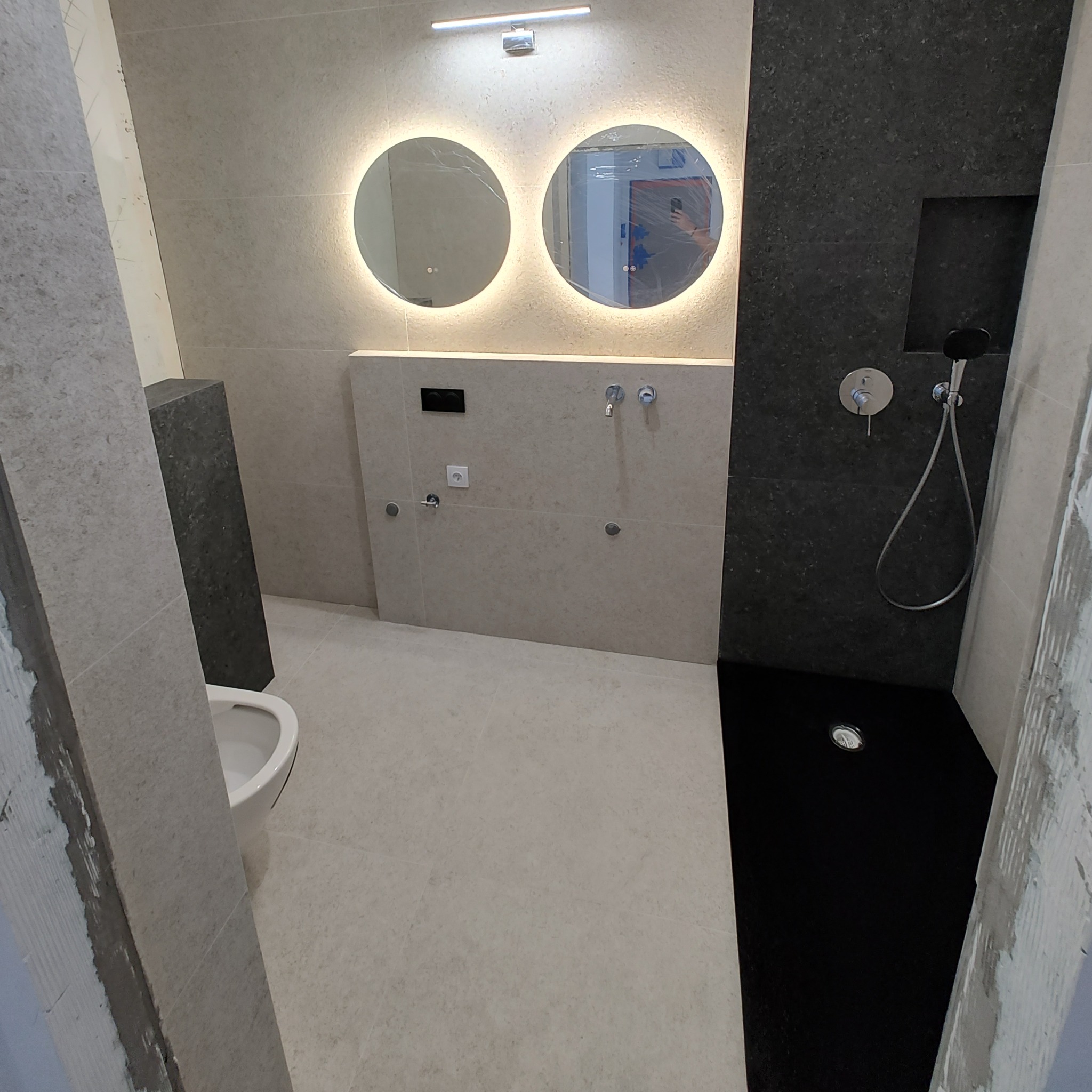 Nowoczesna łazienka po remoncie z dwoma okrągłymi lustrami z podświetleniem LED, czarną kabiną prysznicową typu walk-in i podwieszaną toaletą.