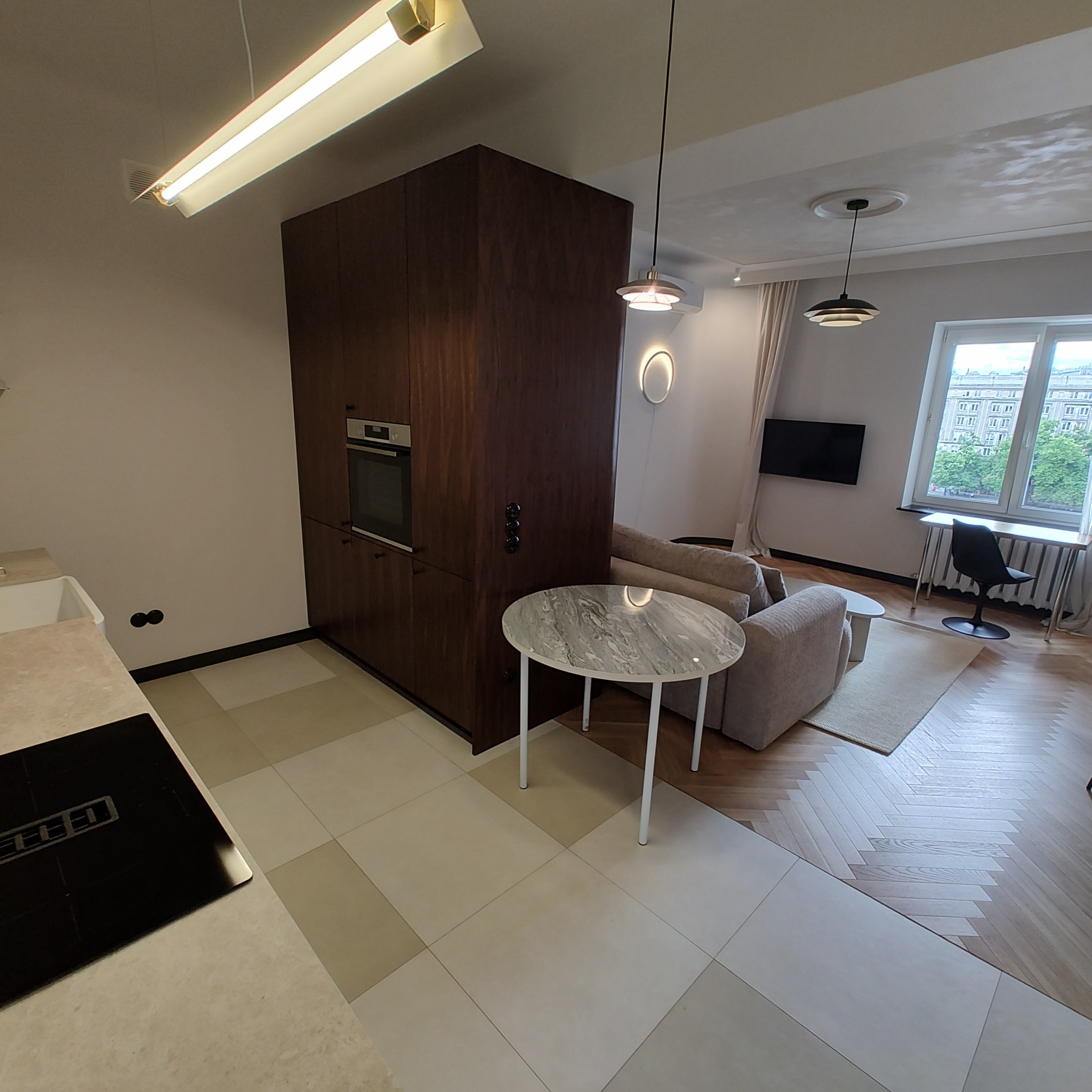 Otwarta przestrzeń mieszkania z widoczną zabudową kuchenną z ciemnego drewna, marmurowym stołem na białych nogach, płytkami podłogowymi w odcieniach beżu i salonem z jasną sofą w tle. W tle...