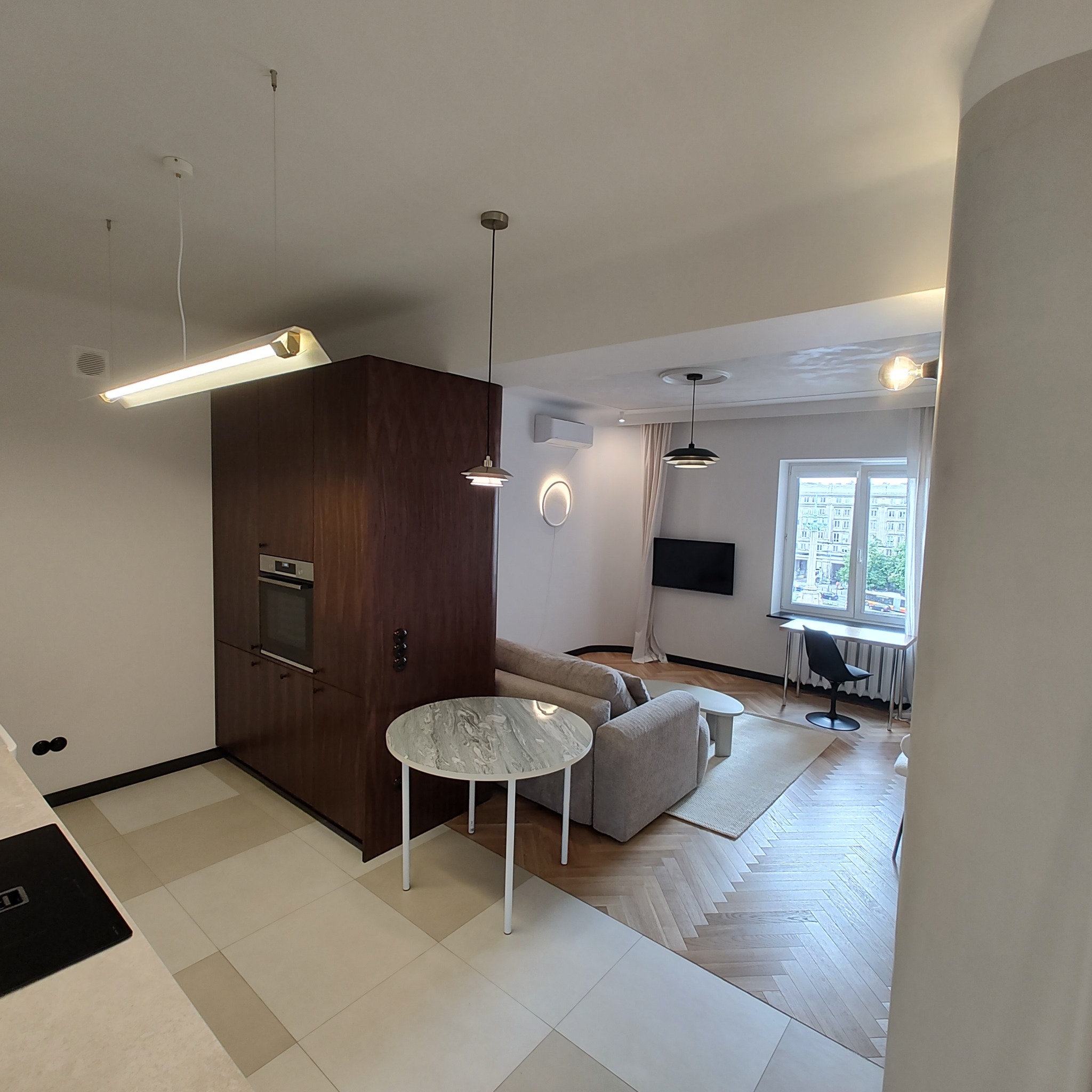 Nowoczesne wnętrze mieszkania z aneksem kuchennym, drewnianą zabudową kuchenną z piekarnikiem, marmurowym stolikiem kawowym, parkietem w jodełkę i widokiem na miasto zza okna.