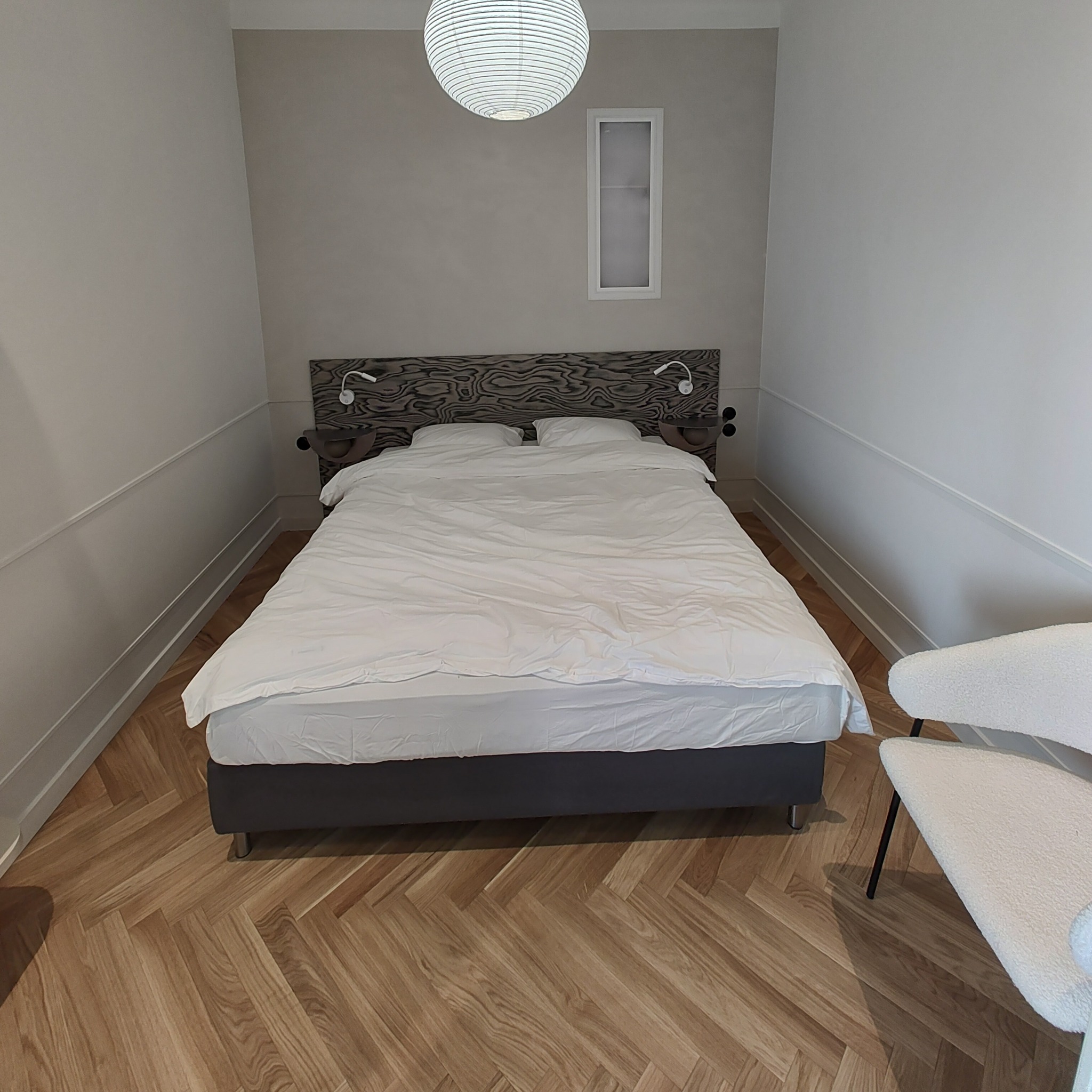 Sypialnia z parkietem ułożonym w jodełkę, łóżkiem z szarym wezgłowiem i białą pościelą, drewnianym panelem nad łóżkiem, oraz designerskim krzesłem obok. Na ścianie lampki nocne i biała wnęka.