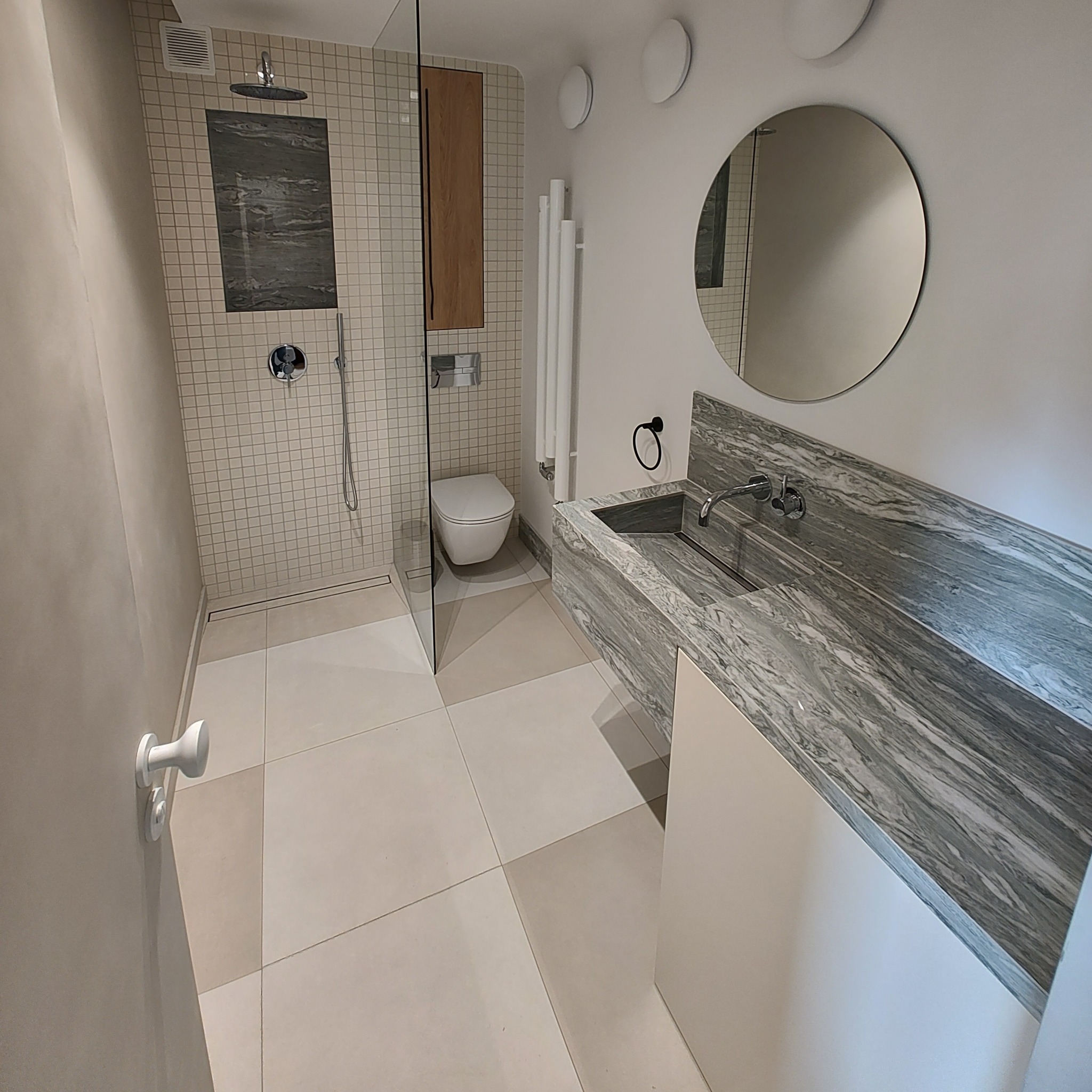 Nowoczesna łazienka z kabiną prysznicową wyłożoną mozaiką, toaletą typu Geberit, grzejnikiem drabinkowym i umywalką z blatem z szarego marmuru.