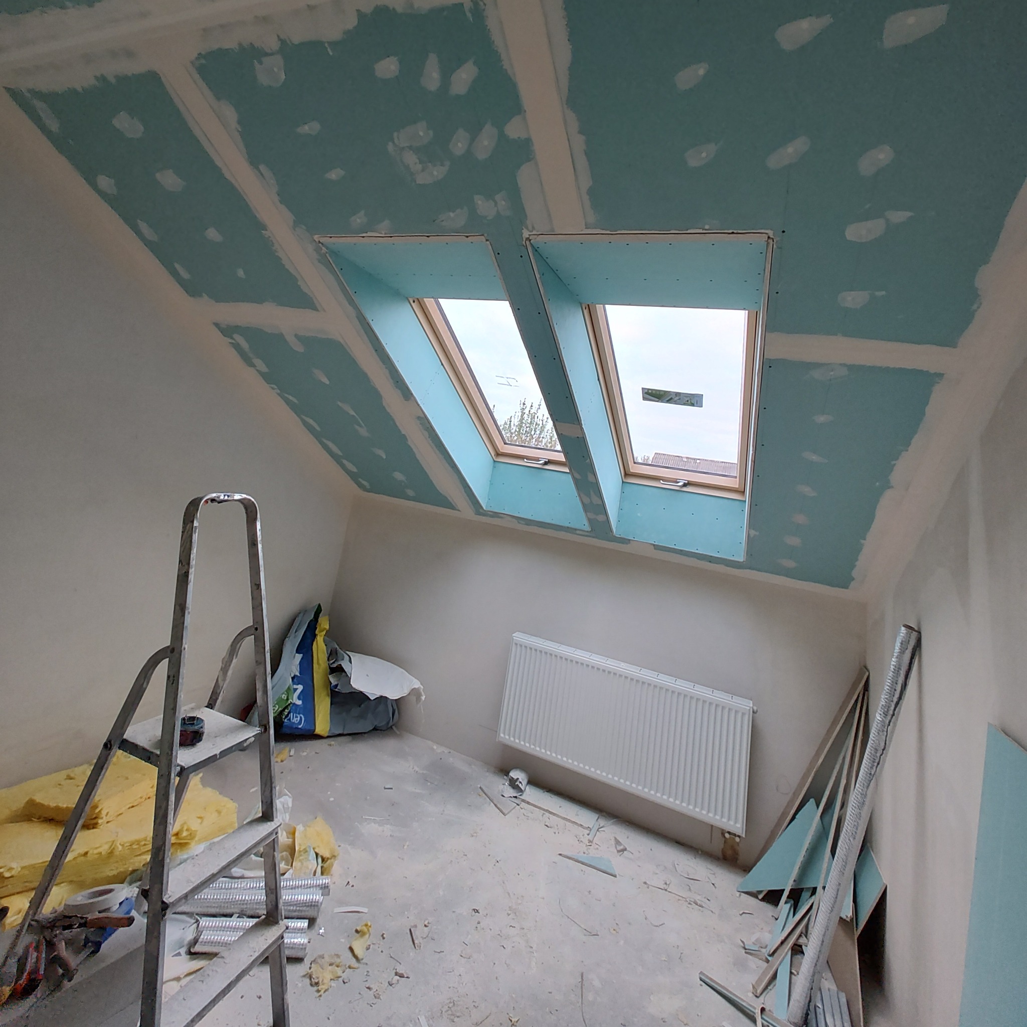 Poddasze w trakcie prac wykończeniowych: zamontowane okna dachowe, płyty gipsowo-kartonowe na suficie z widocznymi łączeniami, drabina, materiały izolacyjne i resztki po budowie na podłodze.