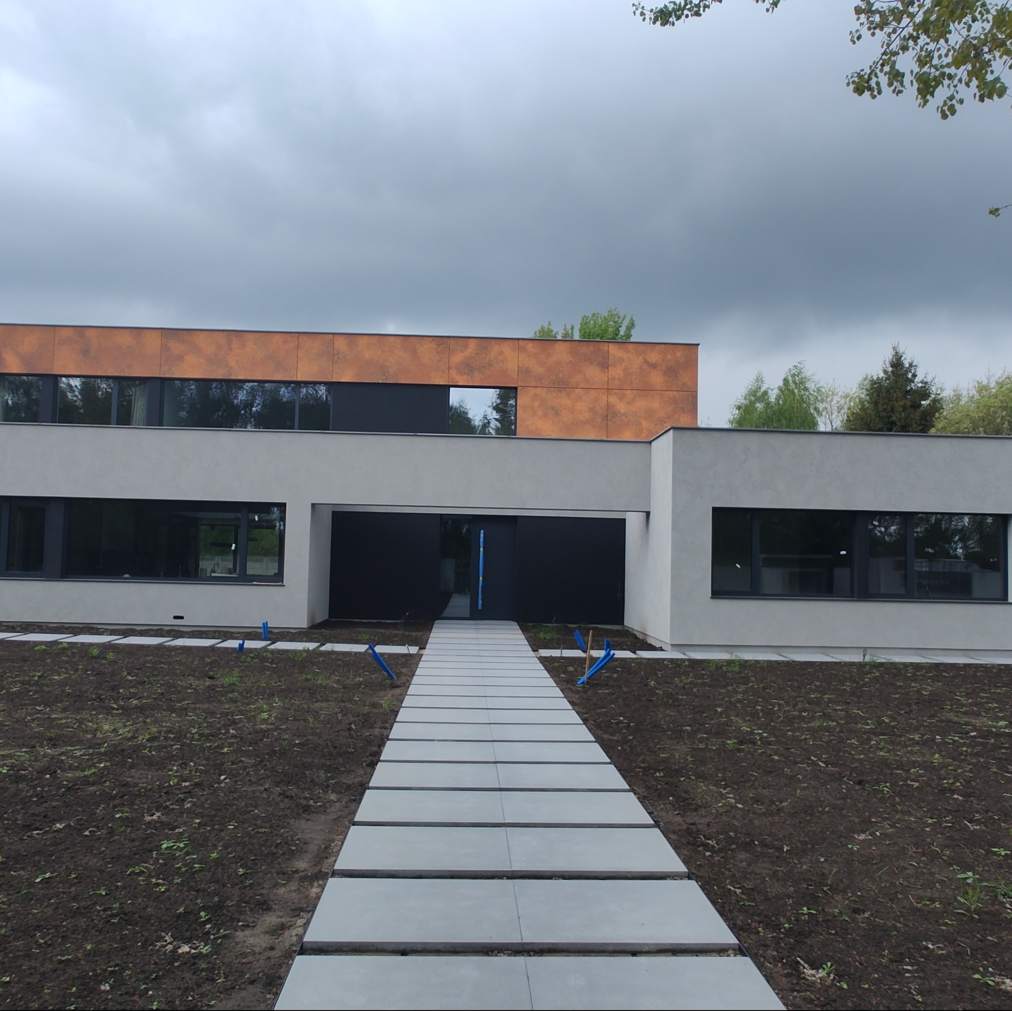 Nowoczesny dom z elewacją w odcieniach szarości i brązu, z minimalistycznym chodnikiem z płyt betonowych prowadzącym do czarnych drzwi wejściowych, otoczony świeżo posadzoną roślinnością.