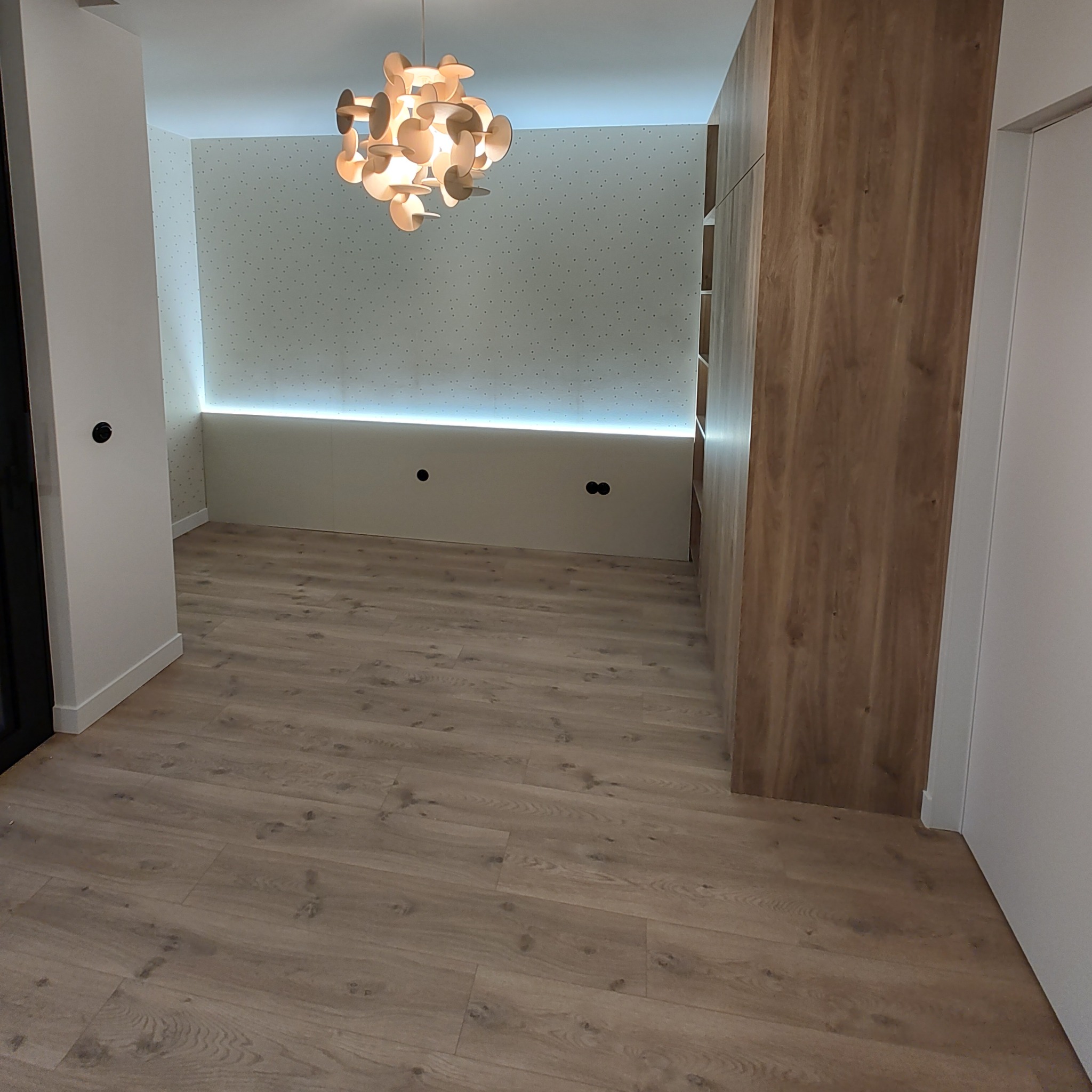 Nowoczesne wnętrze pokoju z drewnianą podłogą, oświetleniem LED pod ścianą i designerską lampą wiszącą, ukazujące minimalistyczny styl i jasne kolory.