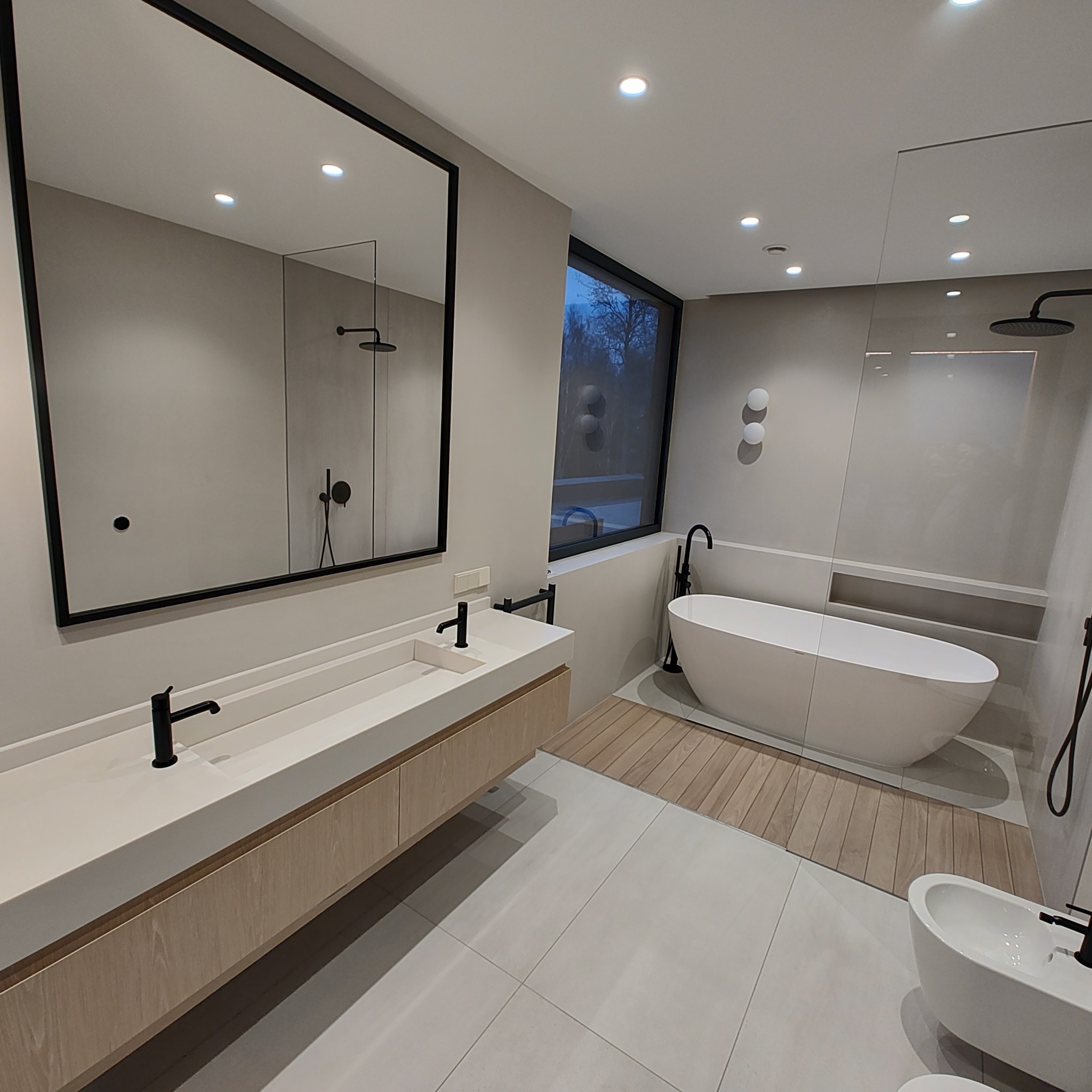 Nowoczesna łazienka z owalną wanną, podwieszaną umywalką z szafką z jasnego drewna, czarną armaturą i dużym lustrem w czarnej ramie.