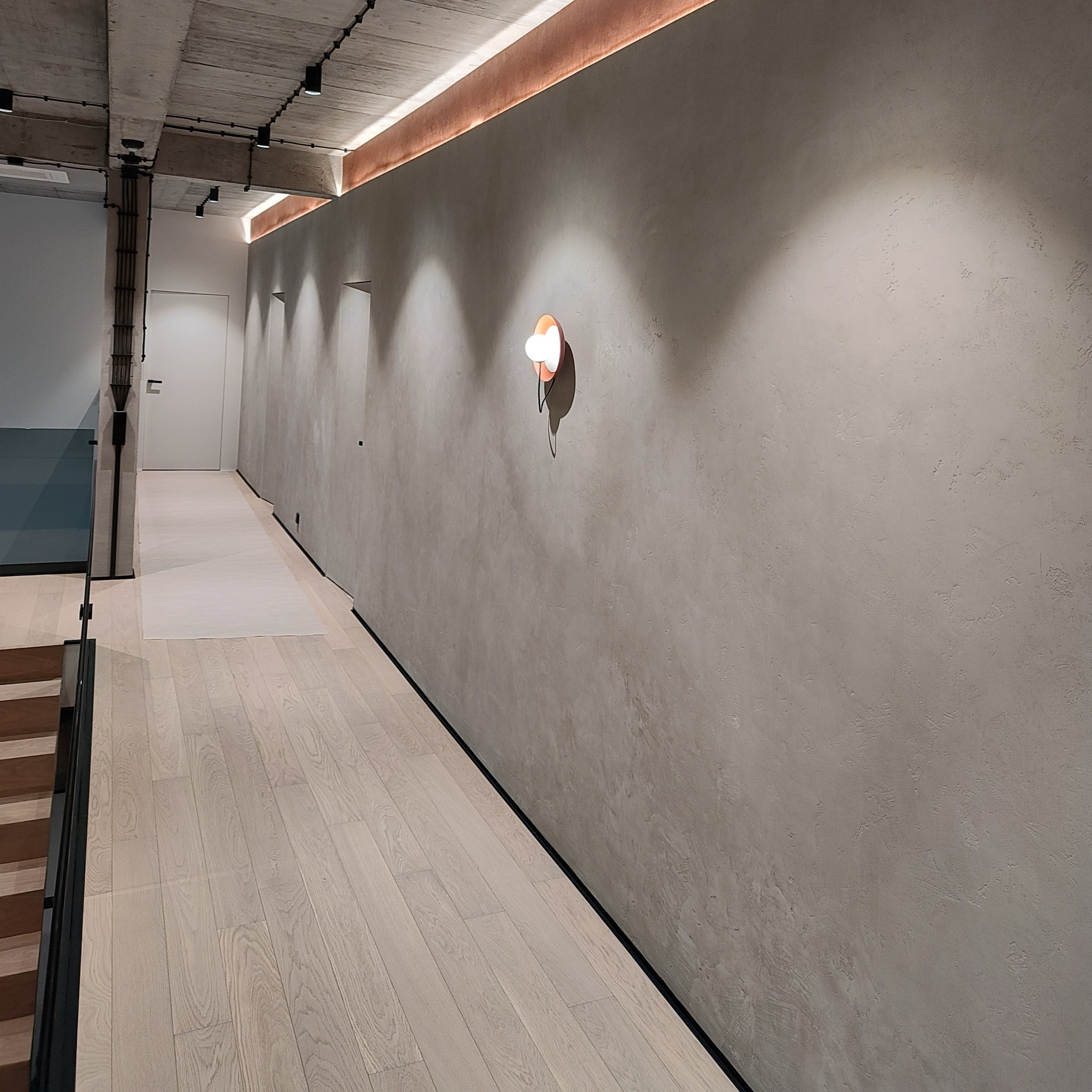 Korytarz z drewnianą podłogą i ścianą wykończoną dekoracyjnym tynkiem w odcieniach szarości, oświetlony listwą LED i kinkietem z okrągłym kloszem.