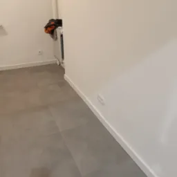 Nowoczesne, matowe, szare płytki podłogowe ułożone w kwadraty, białe ściany z listwami przypodłogowymi i gniazdami elektrycznymi, widoczny grzejnik i rzeczy na nim w tle.