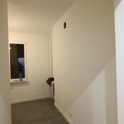 AD-REMONTY I WYKOŃCZENIA ADAM ŻMIJA - Korytarz z szarymi płytkami podłogowymi, świeżo pomalowanymi białymi ścianami, grzejnikiem i oknem, w którym odbija się osoba robiąca zdjęcie.