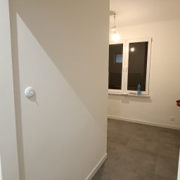 AD-REMONTY I WYKOŃCZENIA ADAM ŻMIJA - Wnętrze mieszkania po odświeżeniu: pomalowane ściany w odcieniach bieli, szara podłoga z płytek, biała listwa przypodłogowa i włącznik światła na ścianie.