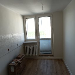 AD-REMONTY I WYKOŃCZENIA ADAM ŻMIJA - Pokój z ułożoną podłogą z paneli, fragment ściany wykończony dekoracyjnymi płytkami, widoczne kartony z materiałami budowlanymi oraz grzejnik pod oknem balkonowym.
