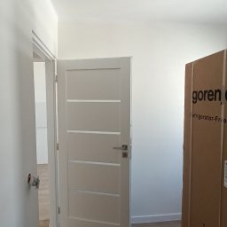 AD-REMONTY I WYKOŃCZENIA ADAM ŻMIJA - Wnętrze w trakcie wykańczania: białe ściany, jasna podłoga, częściowo zamontowane białe drzwi z poziomymi przeszkleniami, wystająca puszka elektryczna i kartonowe opakowanie sprzętu AGD marki Gorenje.