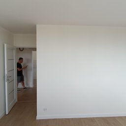 AD-REMONTY I WYKOŃCZENIA ADAM ŻMIJA - Pomalowane na biało ściany w pustym pokoju z drewnianą podłogą, widoczny fragment drugiego pomieszczenia z mężczyzną przeglądającym coś na telefonie.