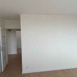 AD-REMONTY I WYKOŃCZENIA ADAM ŻMIJA - Pomalowane na biało wnętrze pokoju z drewnianą podłogą, otwartymi białymi drzwiami wewnętrznymi i widocznym fragmentem instalacji elektrycznej w przejściu do kolejnego pomieszczenia.