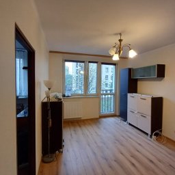 AD-REMONTY I WYKOŃCZENIA ADAM ŻMIJA - Pusty pokój po remoncie z panelami podłogowymi w odcieniach brązu, widoczne meble w zabudowie i lampa stojąca, z oknem balkonowym na zewnątrz widok na blok.