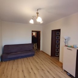 AD-REMONTY I WYKOŃCZENIA ADAM ŻMIJA - Salon po odświeżeniu: świeżo pomalowane ściany w odcieniu złamanej bieli, nowe panele podłogowe w ciepłym odcieniu drewna, widoczna sofa w kolorze ciemnego fioletu, osoba przechodząca przez drzwi...