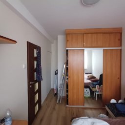 AD-REMONTY I WYKOŃCZENIA ADAM ŻMIJA - Pokój po odświeżeniu: drewniana podłoga, przesuwne drzwi szafy z lustrem odbijającym wnętrze, białe ściany, ciemne drzwi z dekoracyjnymi wstawkami, drabina oparta o ścianę.