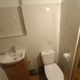 Jasna łazienka po remoncie: białe płytki imitujące marmur na ścianach, drewniana szafka pod umywalką i toaleta. Widoczne detale instalacji.