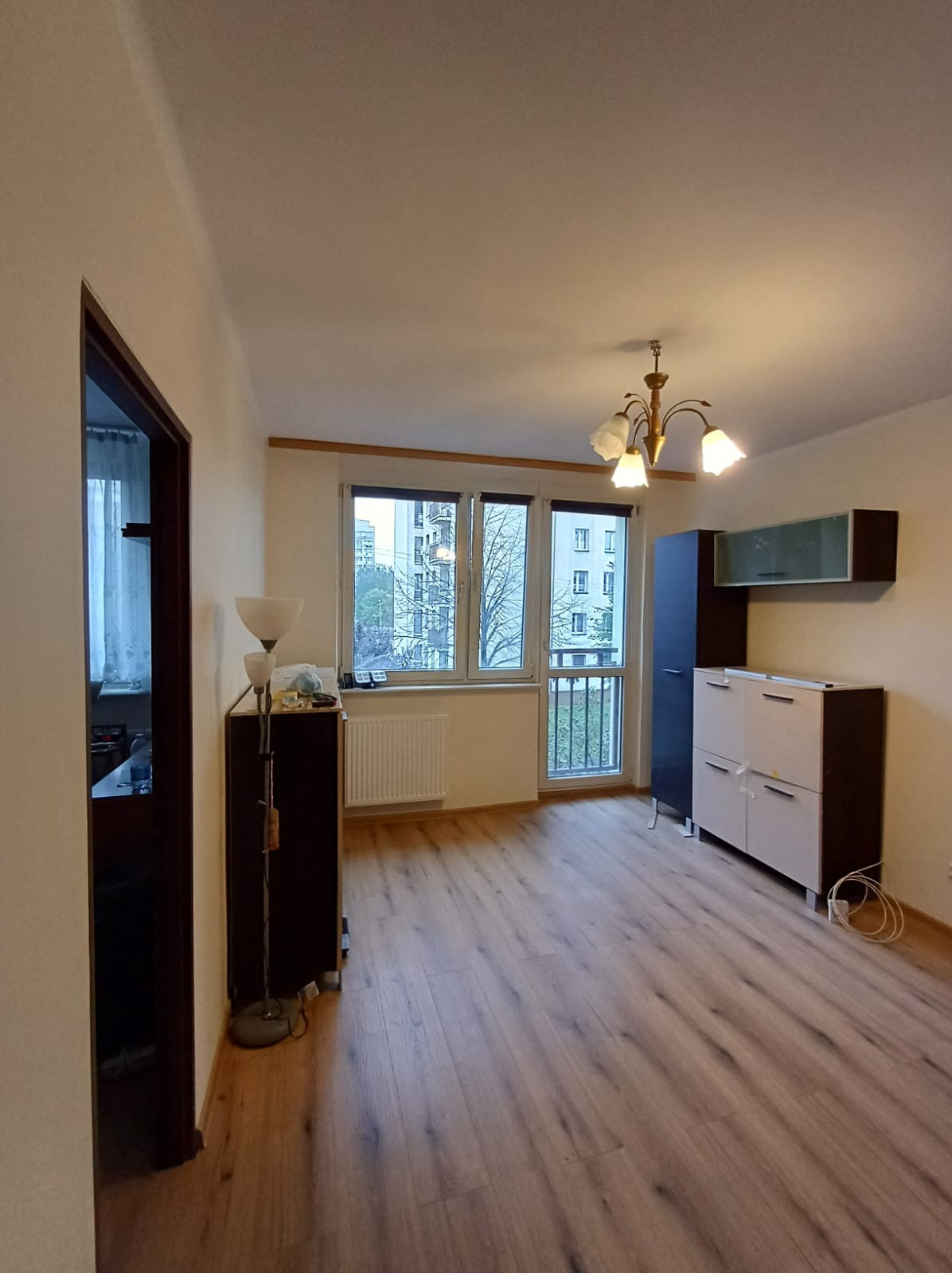 Pusty pokój po remoncie z panelami podłogowymi w odcieniach brązu, widoczne meble w zabudowie i lampa stojąca, z oknem balkonowym na zewnątrz widok na blok.