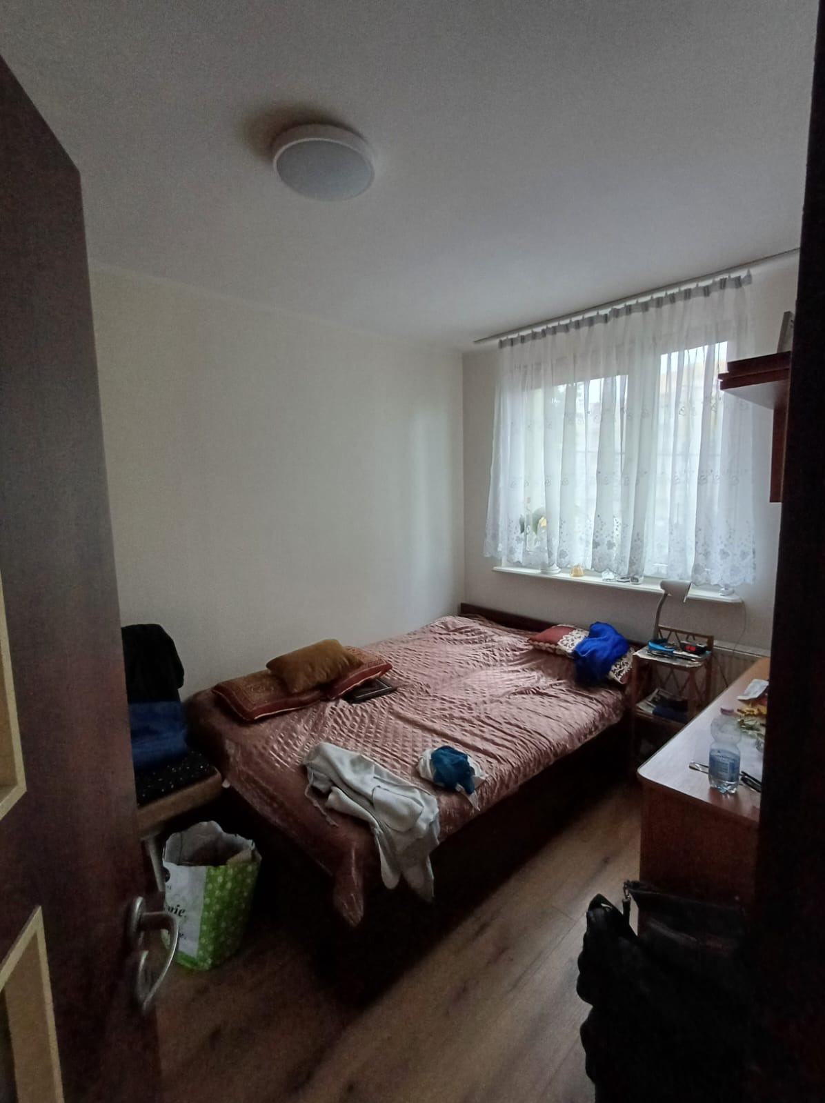 Pokój z łóżkiem, biurkiem i oknem z białymi zasłonami, widok z perspektywy drzwi, widoczny kosz na śmieci i torba na podłodze.