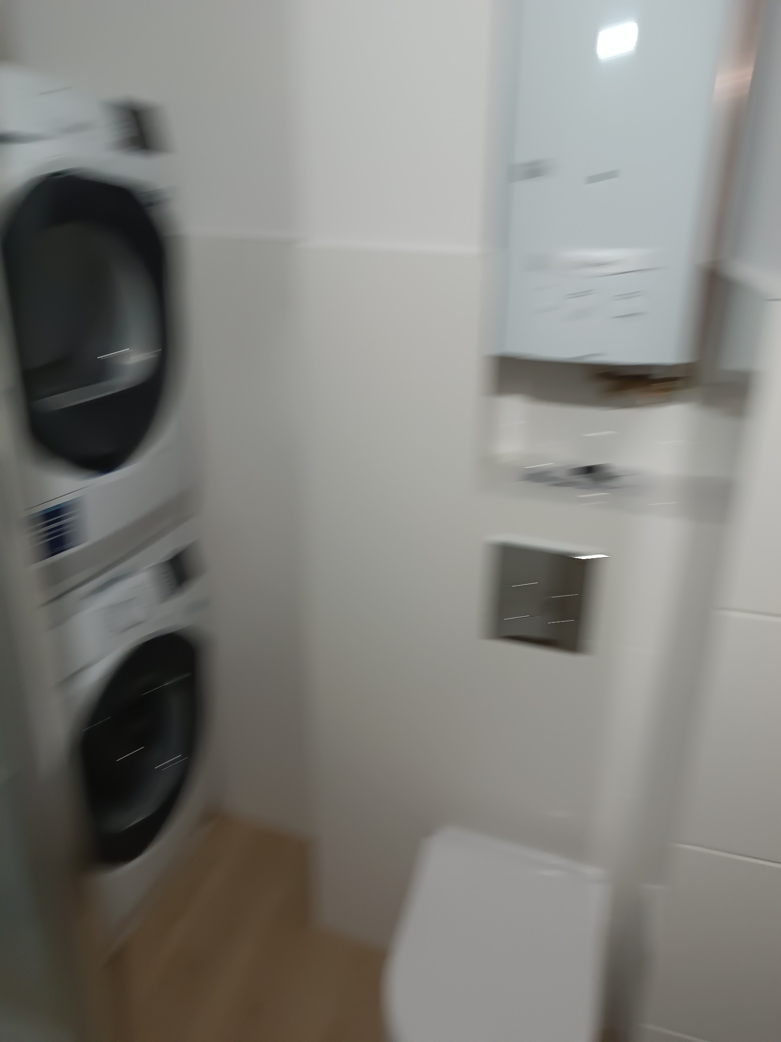 Biała łazienka z pralką i suszarką w słupku, wiszącą toaletą oraz piecykiem gazowym. Minimalistyczny styl, jasne wnętrze.