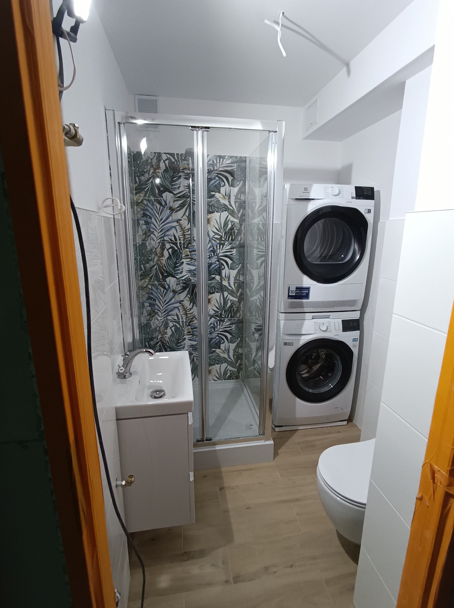 Nowoczesna łazienka po remoncie: przeszklona kabina prysznicowa z motywem roślinnym, umywalka z szafką, toaleta i pralko-suszarka w kolumnie Electrolux.