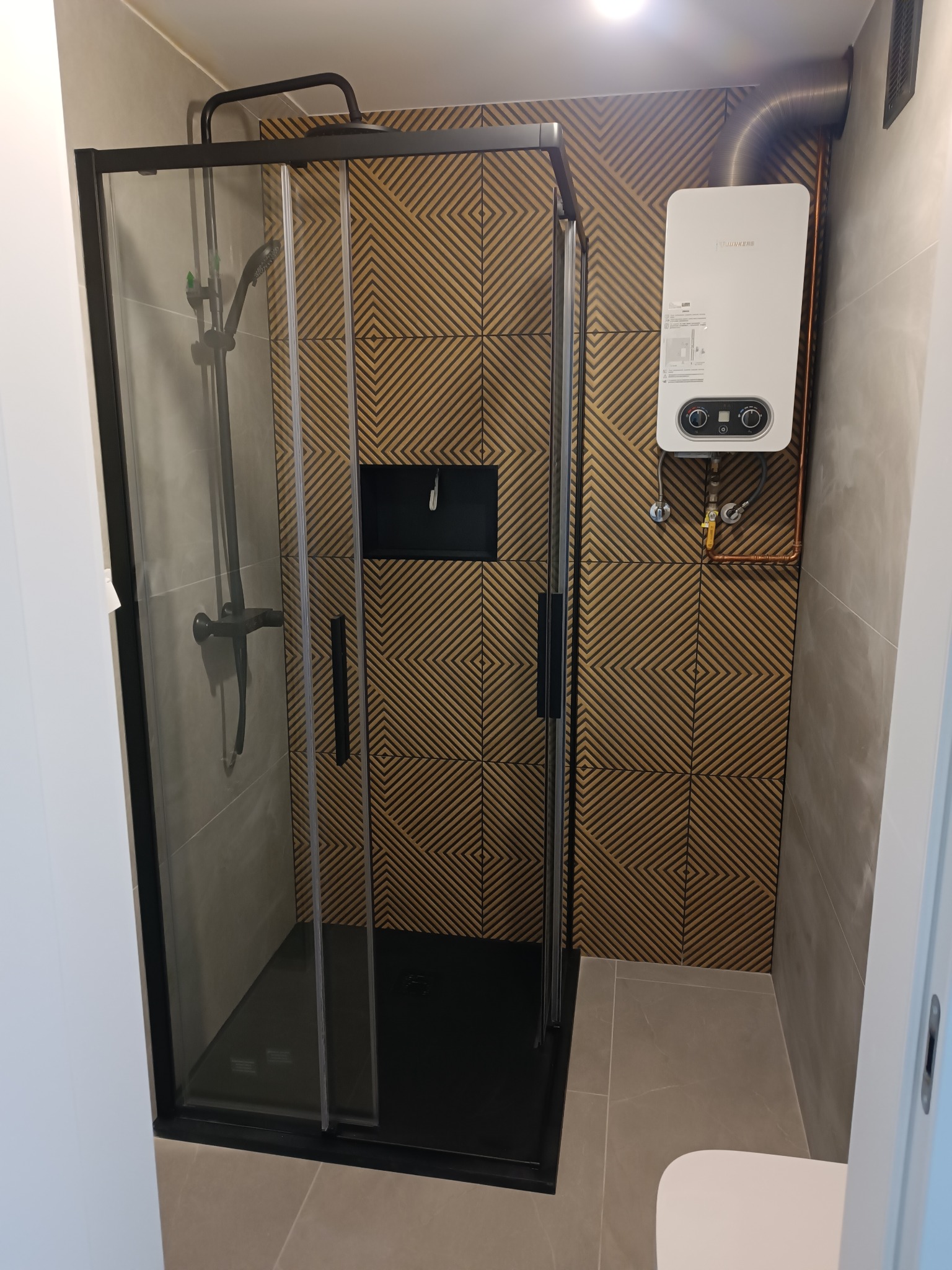 Nowoczesna kabina prysznicowa z czarną armaturą i geometrycznymi płytkami na ścianie w odcieniach brązu. Zamontowany bojler i szara podłoga. Wykończona łazienka.