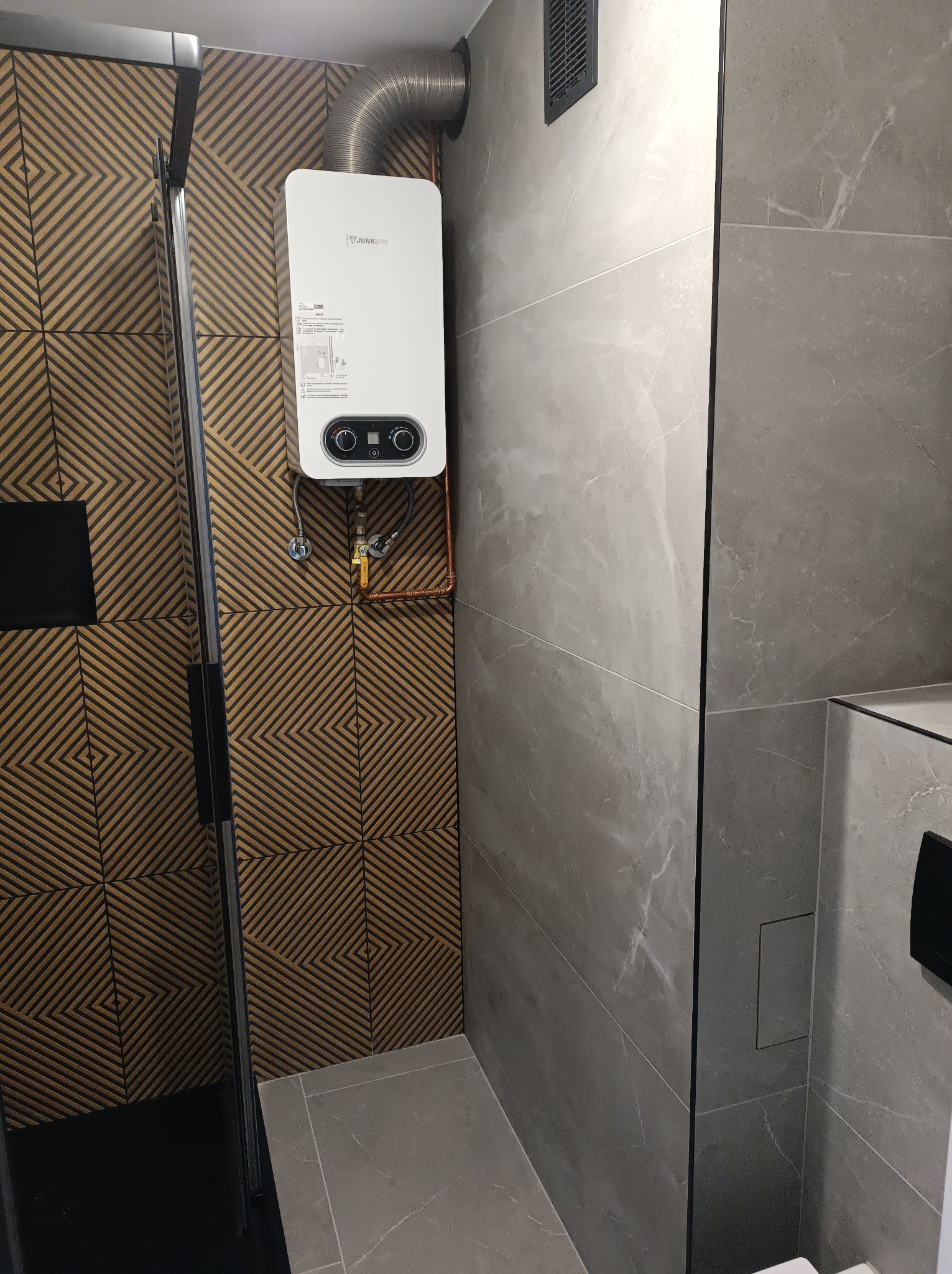 Nowoczesna łazienka z płytkami w geometryczny wzór i marmurkową teksturą. Widoczny biały piec gazowy Junkers z metalową rurą, czarna kabina prysznicowa.