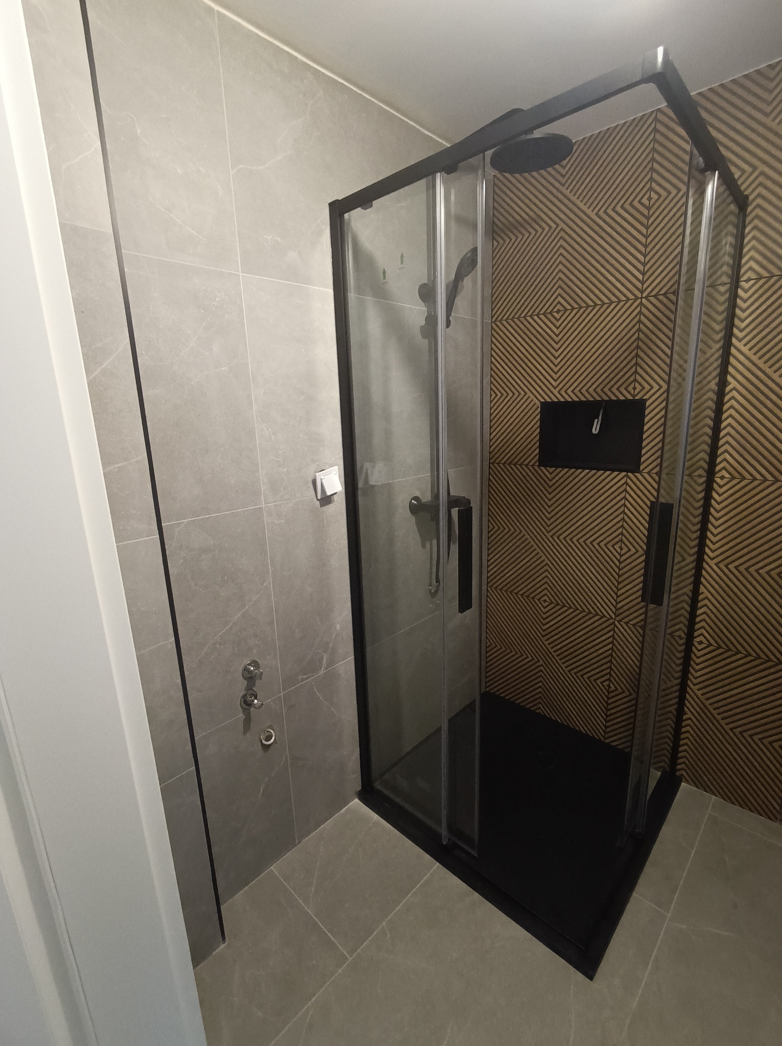 Nowoczesna kabina prysznicowa z czarną ramą i deszczownicą, na tle płytek w geometryczny wzór. Szare płytki na ścianach i podłodze. Minimalistyczny design.