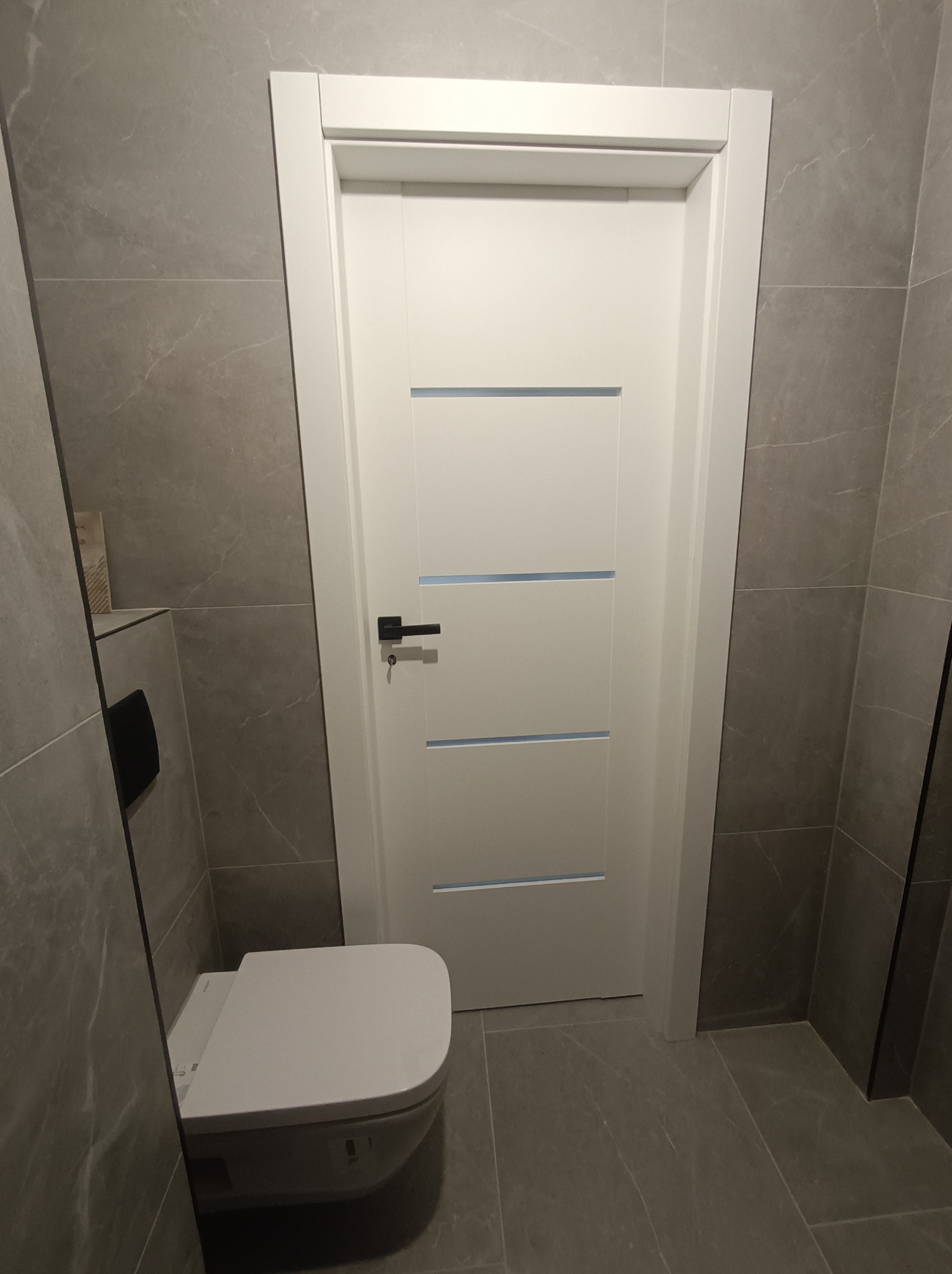 Nowoczesna łazienka z szarymi płytkami, białą, minimalistyczną toaletą i drzwiami z poziomymi przeszkleniami, czarna klamka. Całość utrzymana w stonowanej kolorystyce.