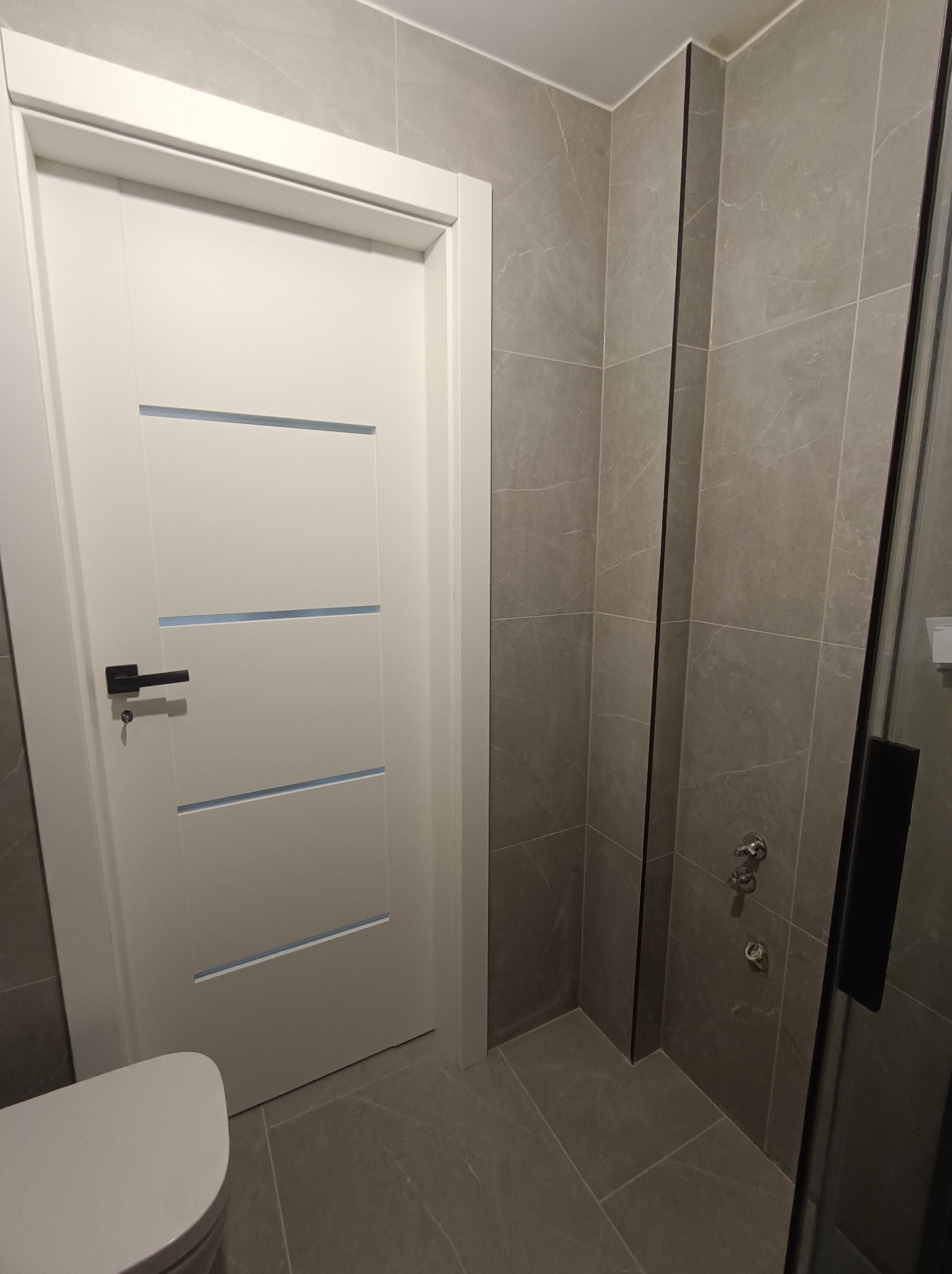 Nowoczesna łazienka z szarymi płytkami, białą, minimalistyczną  drzwiami z poziomymi wgłębieniami i czarną klamką. Widoczny fragment toalety.