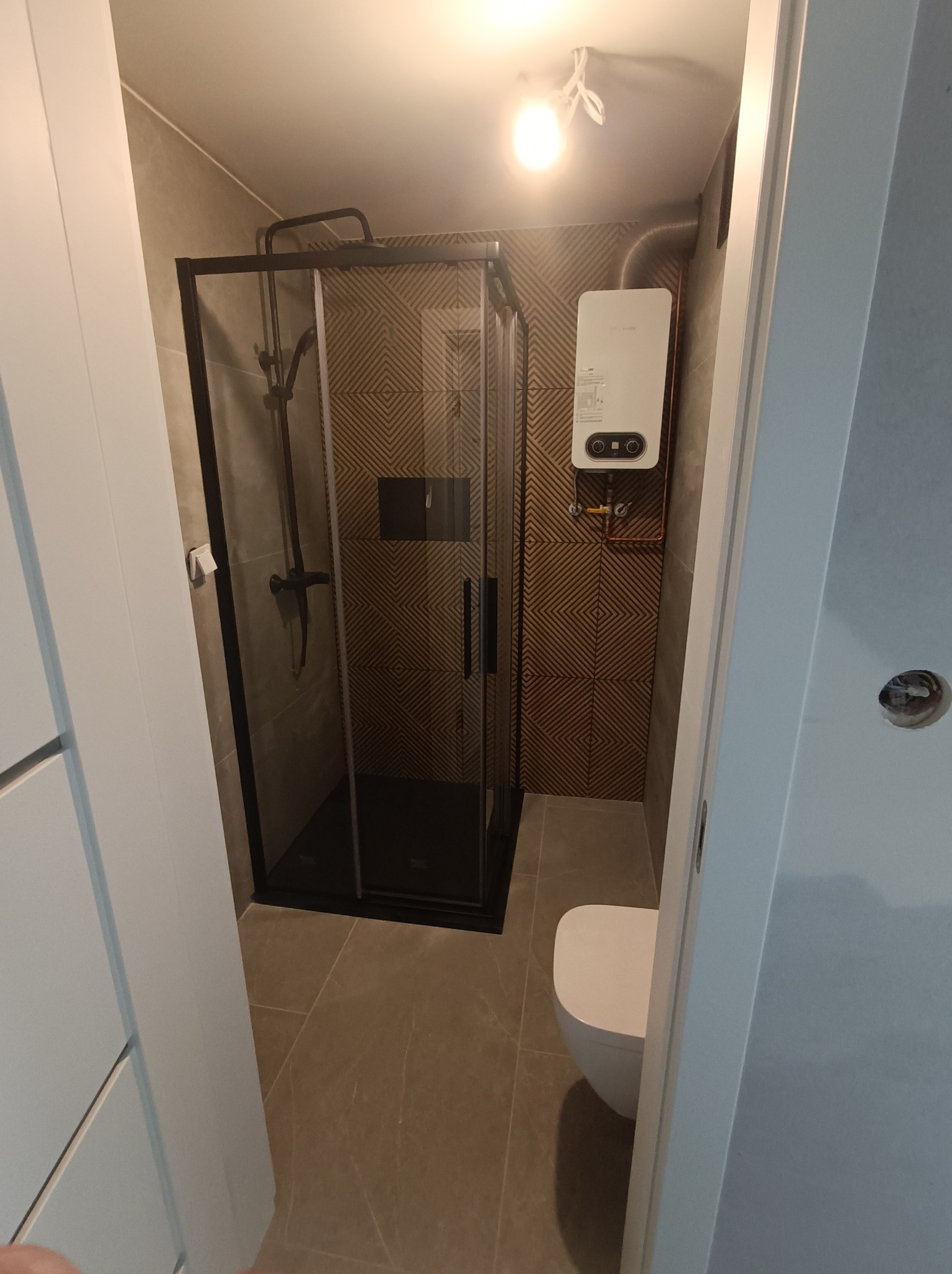 Nowoczesna łazienka po renowacji: kabina prysznicowa z czarną ramą, wzorzyste płytki na ścianie, podwieszany bojler. Minimalistyczny design, widok zza uchylonych drzwi.