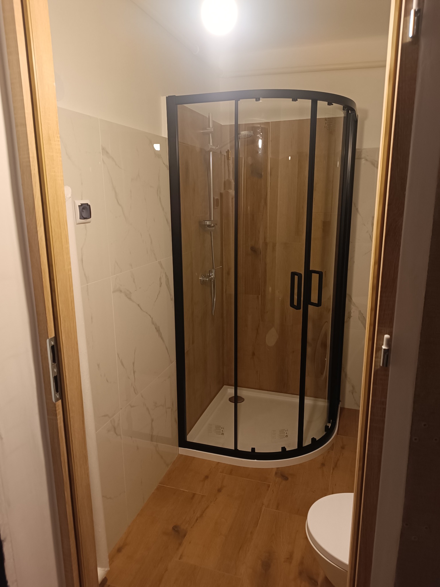 Nowoczesna kabina prysznicowa z czarnymi profilami w wykończonej łazience. Widoczne płytki imitujące marmur na ścianach i drewnopodobne na podłodze. Jasne oświetlenie.