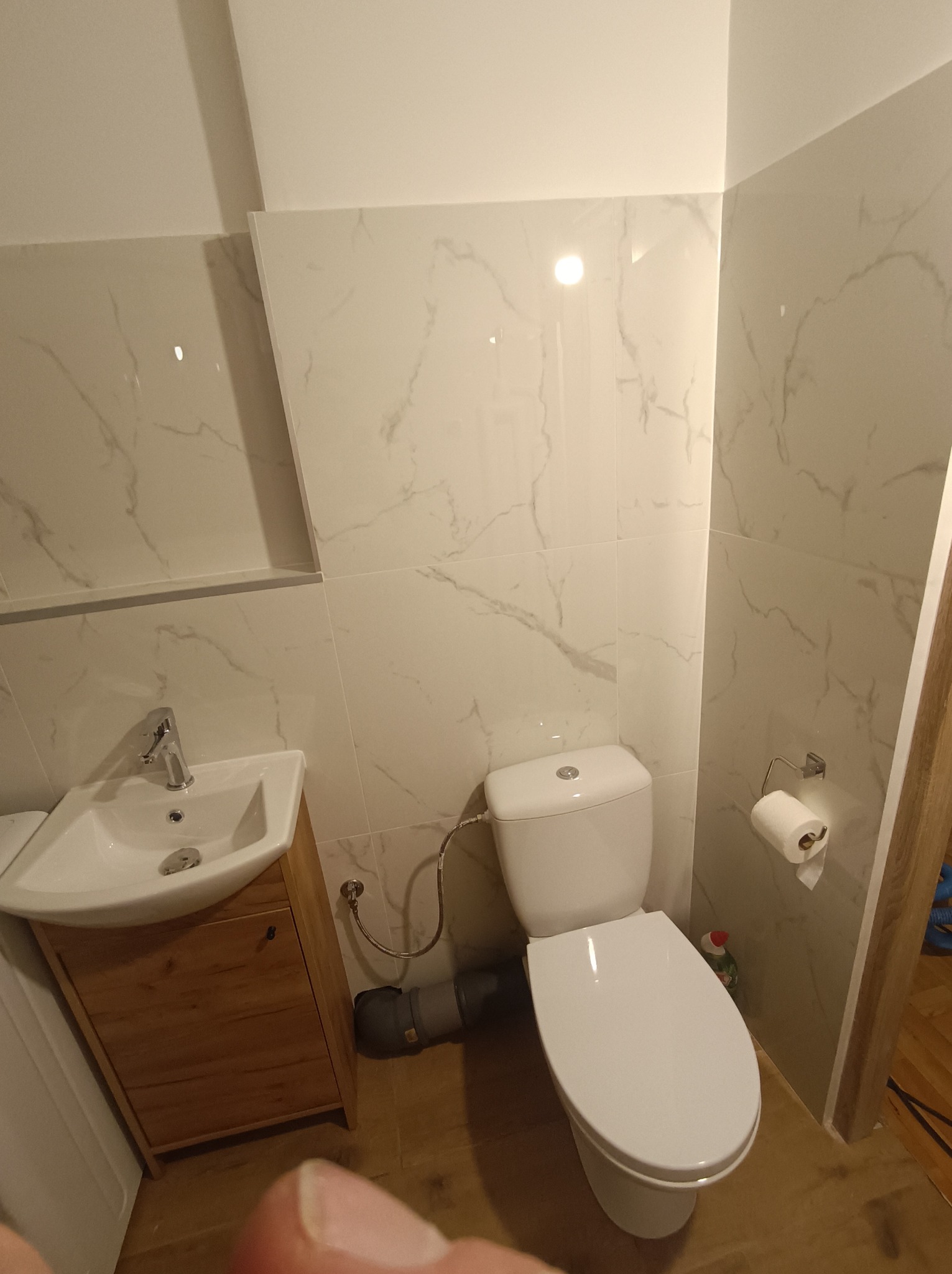 Jasna łazienka po remoncie: białe płytki imitujące marmur na ścianach, drewniana szafka pod umywalką i toaleta. Widoczne detale instalacji.