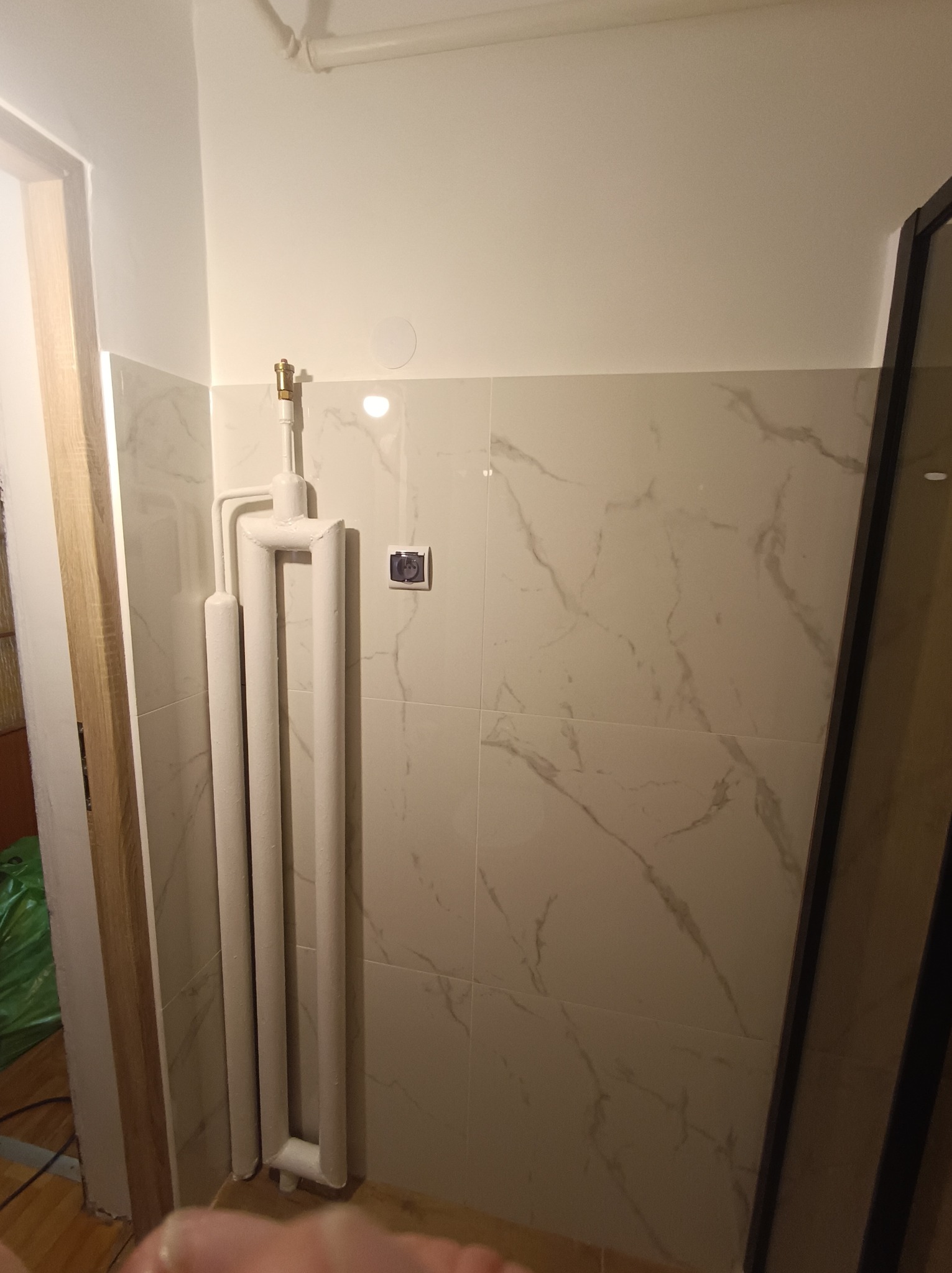 Biały grzejnik drabinkowy na tle ściany z płytek imitujących marmur w wyremontowanej łazience. Widoczny fragment kabiny prysznicowej i drzwi.