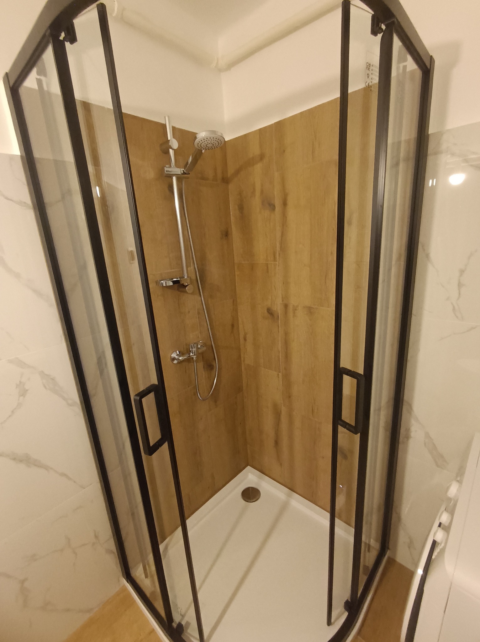 Nowoczesna kabina prysznicowa z czarnymi profilami i drewnopodobnymi płytkami wewnątrz, na tle marmurowej ściany. Widoczny biały brodzik i chromowana armatura.