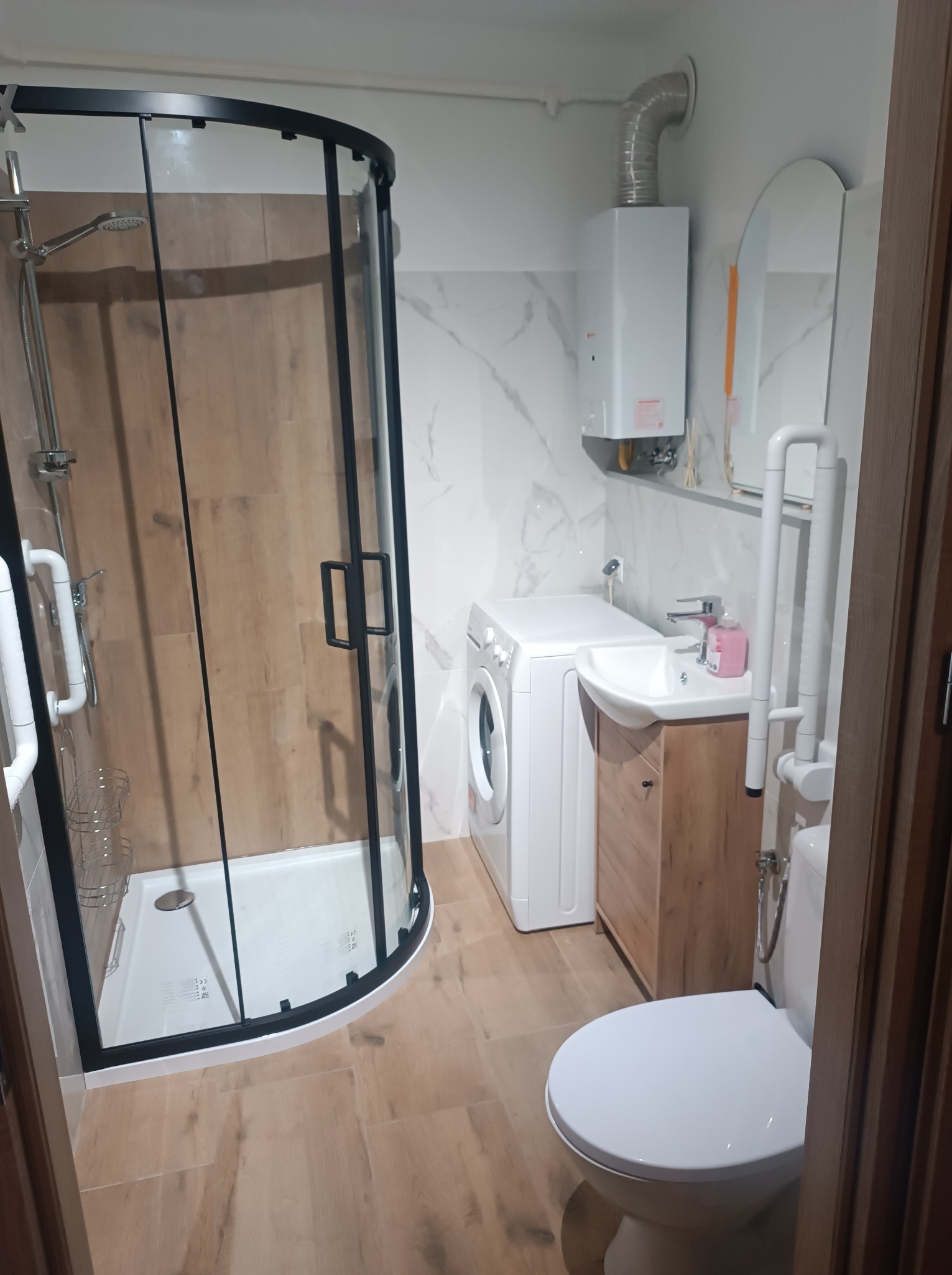 Nowoczesna łazienka po remoncie: kabina prysznicowa z czarną ramą, pralka, umywalka z szafką w odcieniach drewna, toaleta z uchwytami dla niepełnosprawnych i bojler.