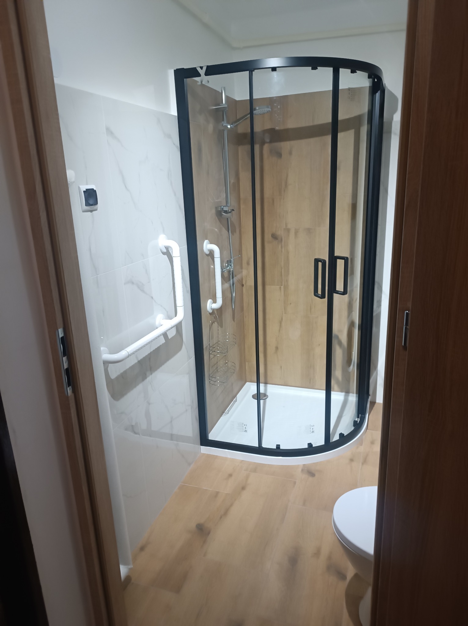 Wykończona łazienka z kabiną prysznicową w czarnej ramie, drewnopodobnymi płytkami i uchwytami dla niepełnosprawnych. Widok zza futryny drzwi.