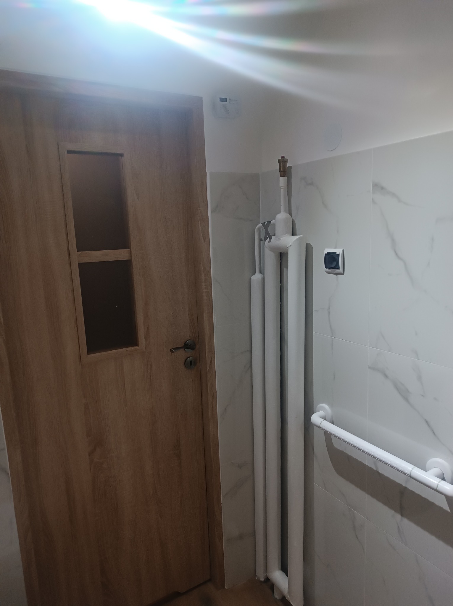 Wykończona łazienka z grzejnikiem drabinkowym, uchwytem dla niepełnosprawnych i drzwiami z przeszkleniem. Białe marmurowe płytki na ścianach.