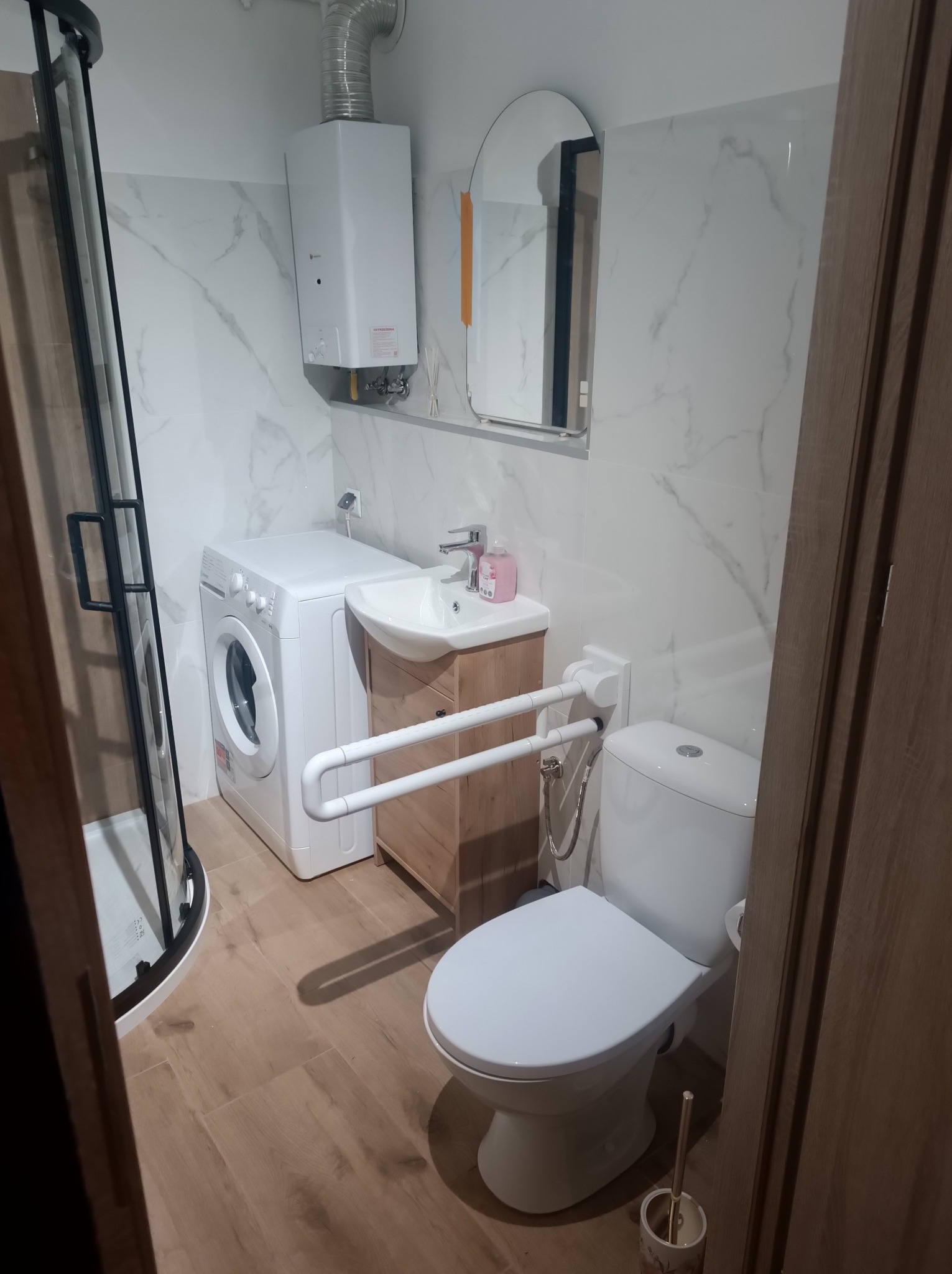 Wykończona łazienka z kabiną prysznicową, pralką, umywalką z szafką i toaletą z uchwytami dla niepełnosprawnych. Białe marmurowe płytki ścienne i drewnopodobna podłoga.